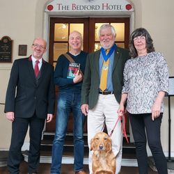 Four adults and a dog standing outside The Bedford Hotel entrance, one person holding a book.