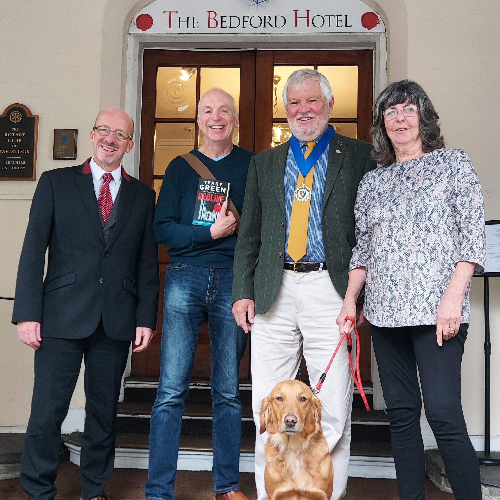 Four adults and a dog standing outside The Bedford Hotel entrance, one person holding a book.