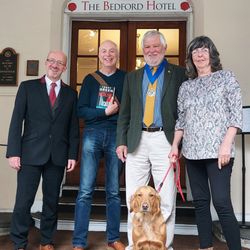 Four adults and a dog standing outside The Bedford Hotel entrance, one person holding a book.