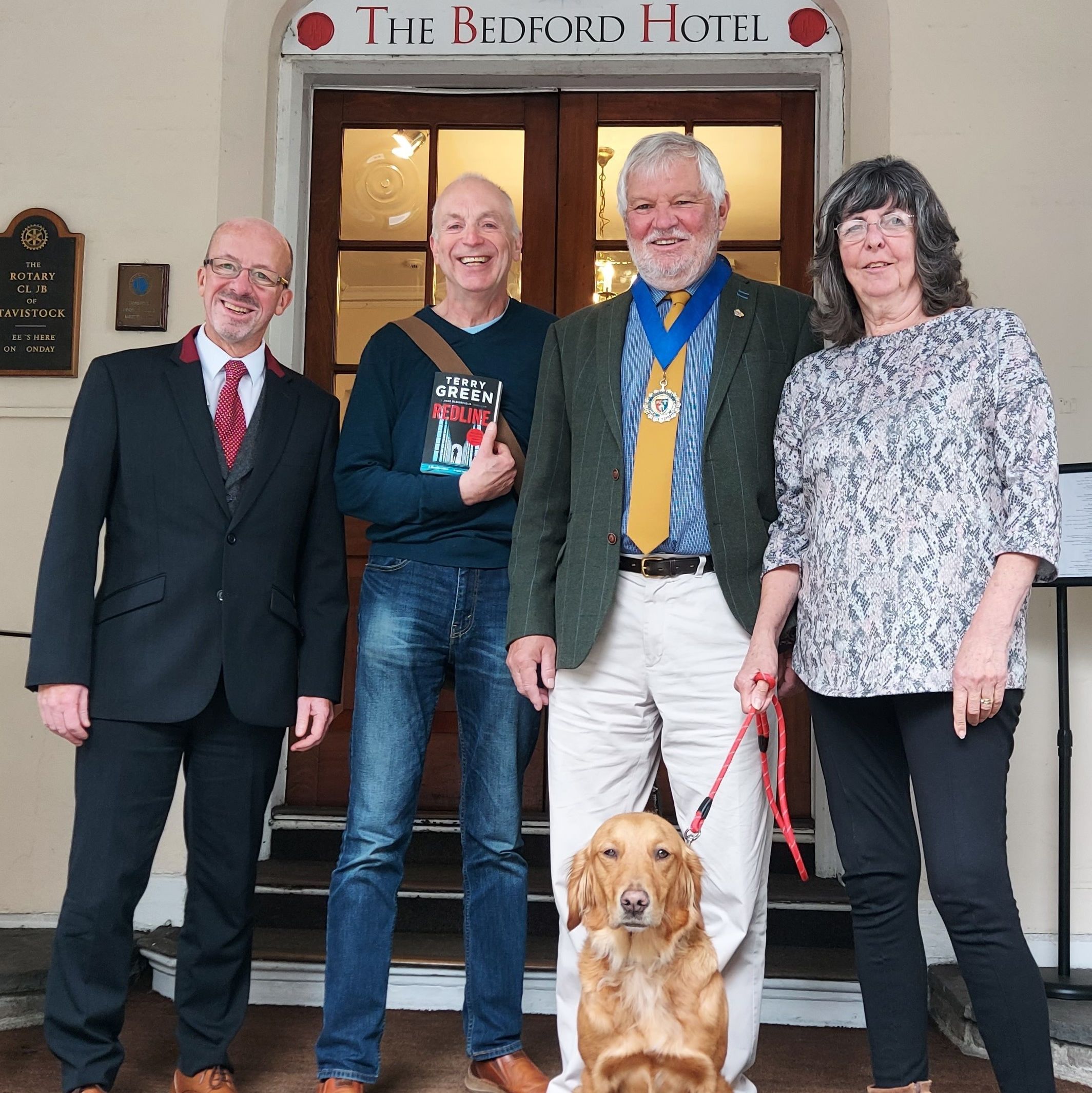 Four adults and a dog standing outside The Bedford Hotel entrance, one person holding a book.