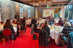 Group of people sitting at round tables having breakfast in a conference room with a projector screen displaying 'Tavistock Plan'.