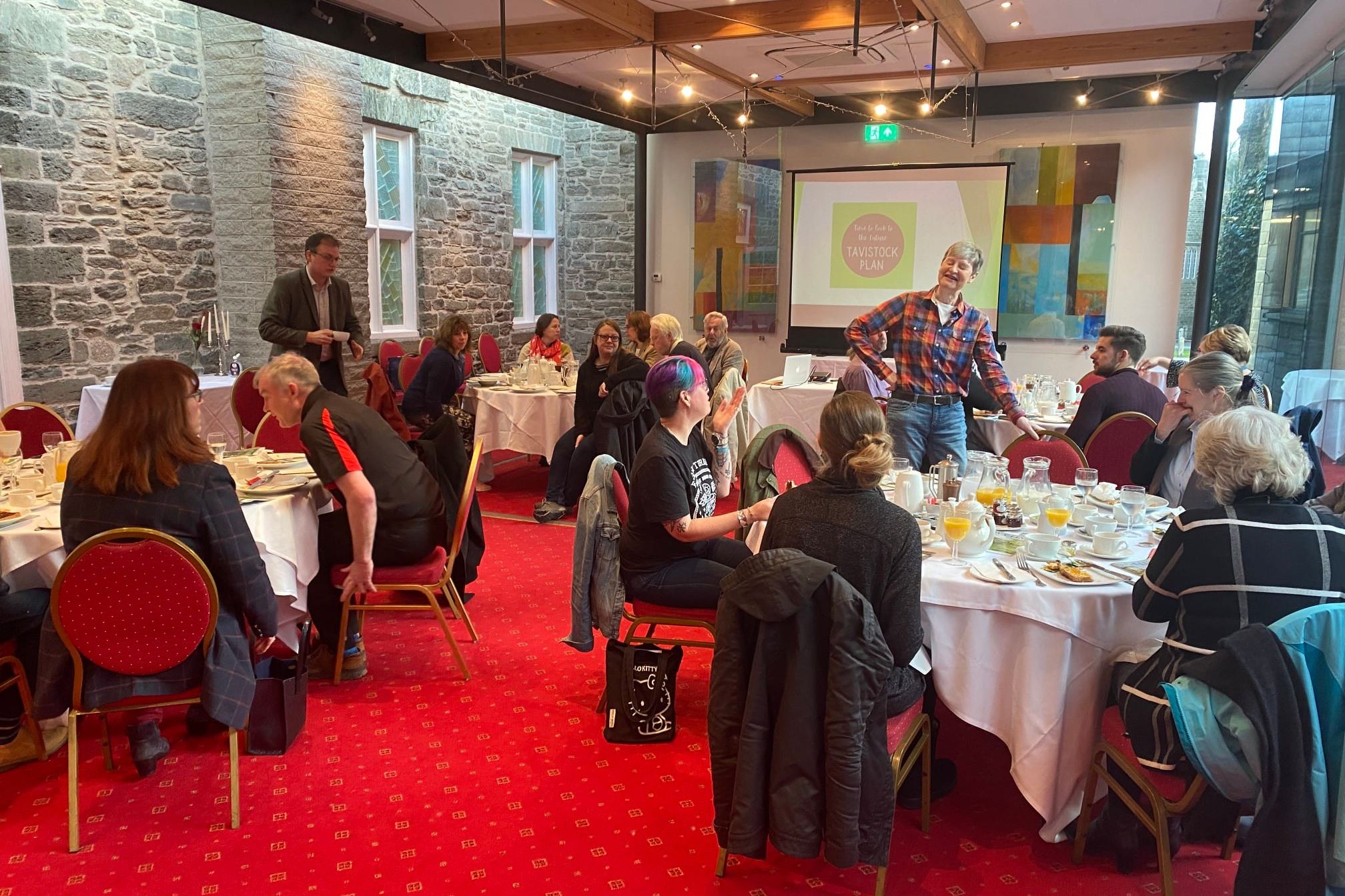 Group of people sitting at round tables having breakfast in a conference room with a projector screen displaying 'Tavistock Plan'.
