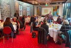 Group of people sitting at round tables having breakfast in a conference room with a projector screen displaying 'Tavistock Plan'.