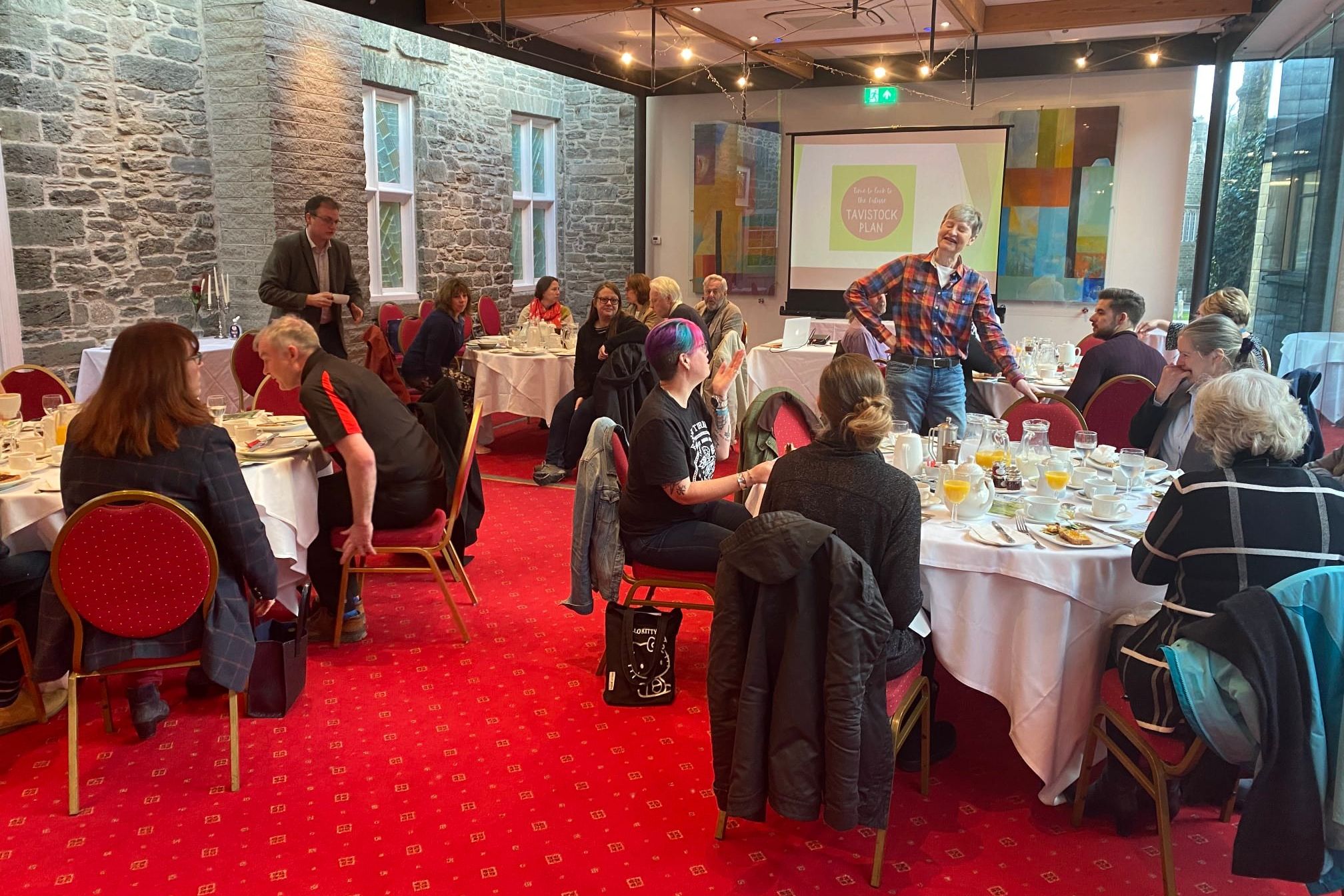 Group of people sitting at round tables having breakfast in a conference room with a projector screen displaying 'Tavistock Plan'.