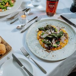 A fine dining table setting with fresh bread rolls, salad, a main course topped with greens, and a glass of ale.
