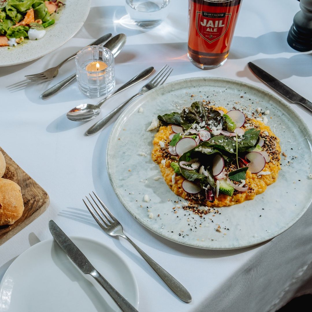 A fine dining table setting with fresh bread rolls, salad, a main course topped with greens, and a glass of ale.