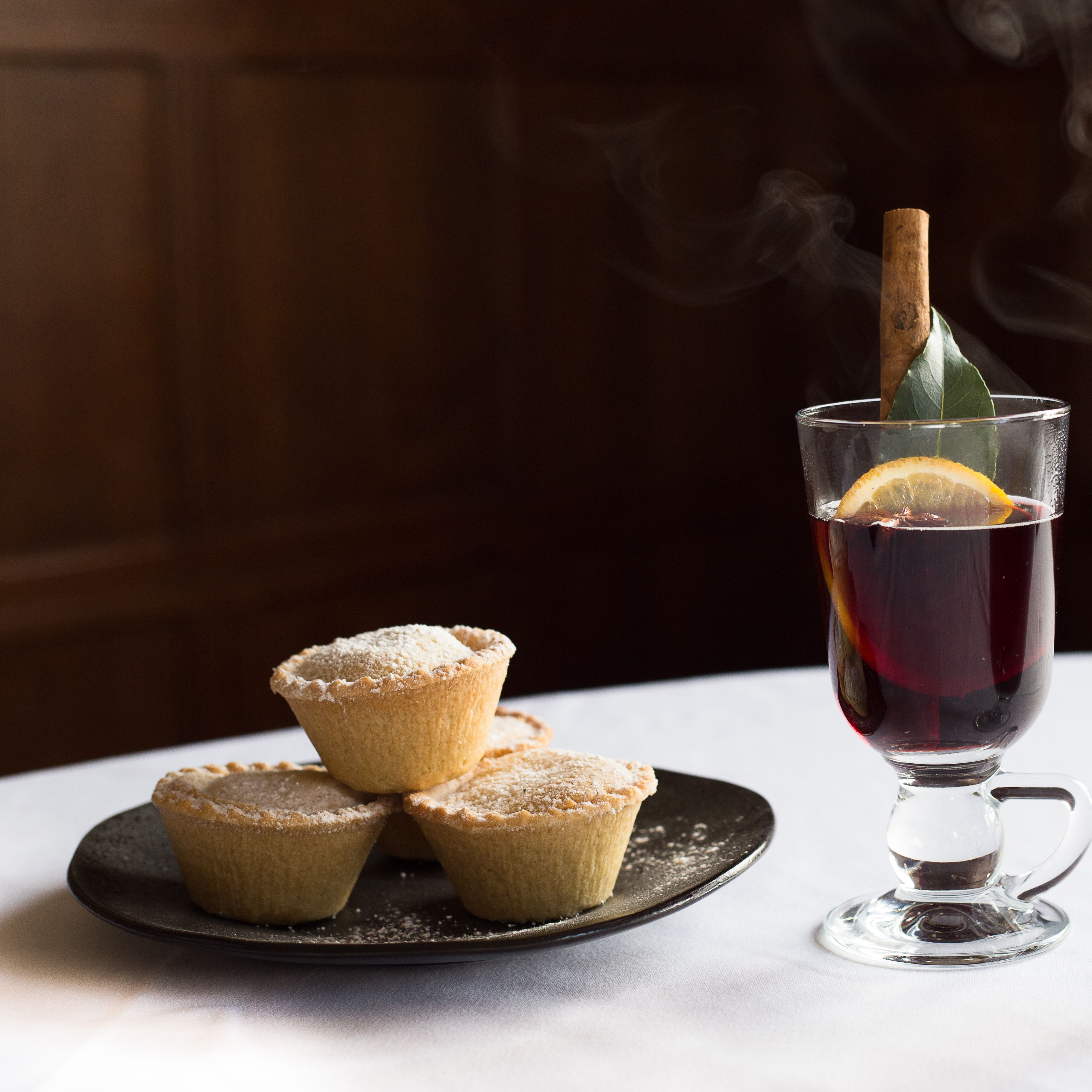 Plate of mince pies beside two glasses of mulled wine garnished with orange, bay leaf, and cinnamon sticks on a white tablecloth
