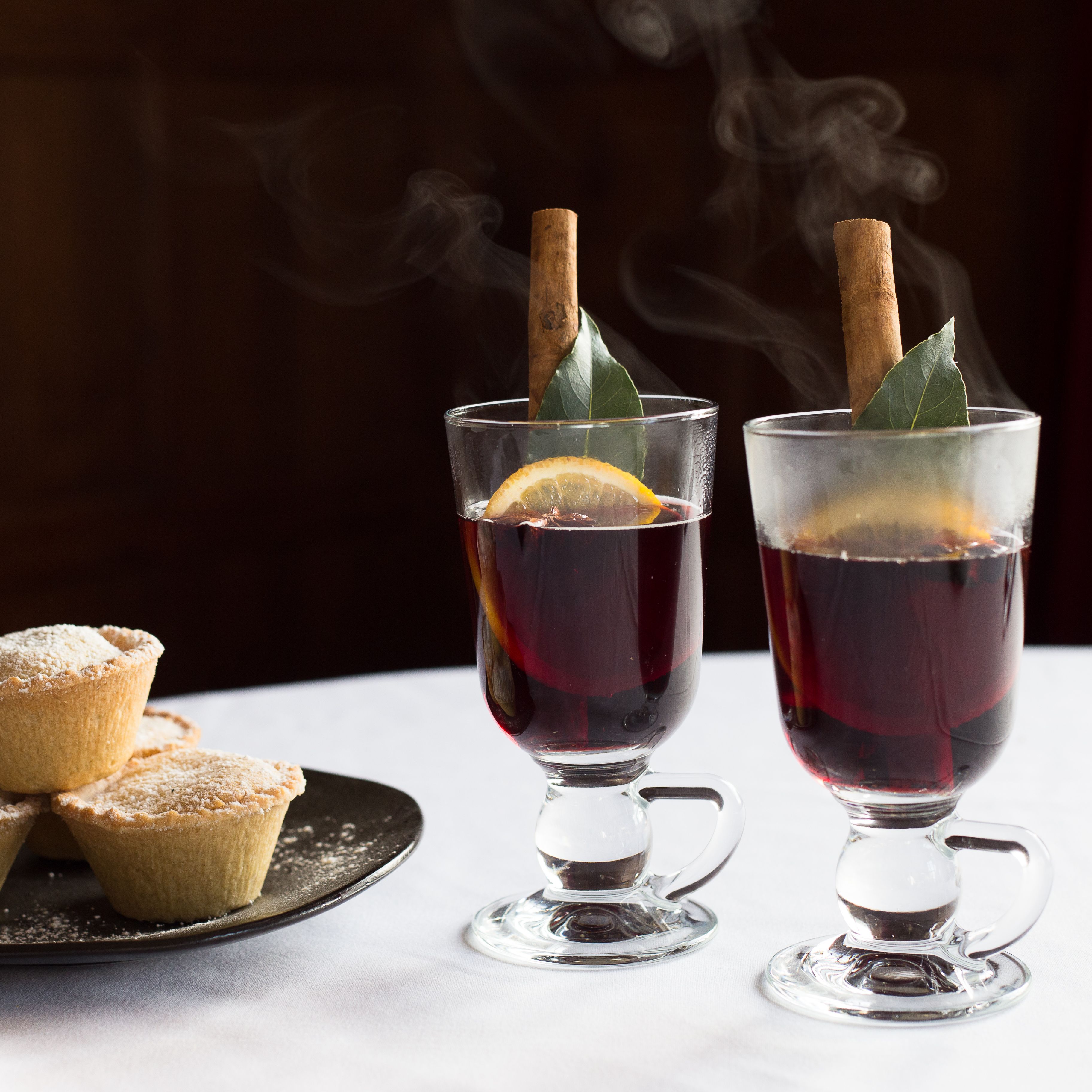 Plate of mince pies beside two glasses of mulled wine garnished with orange, bay leaf, and cinnamon sticks on a white tablecloth