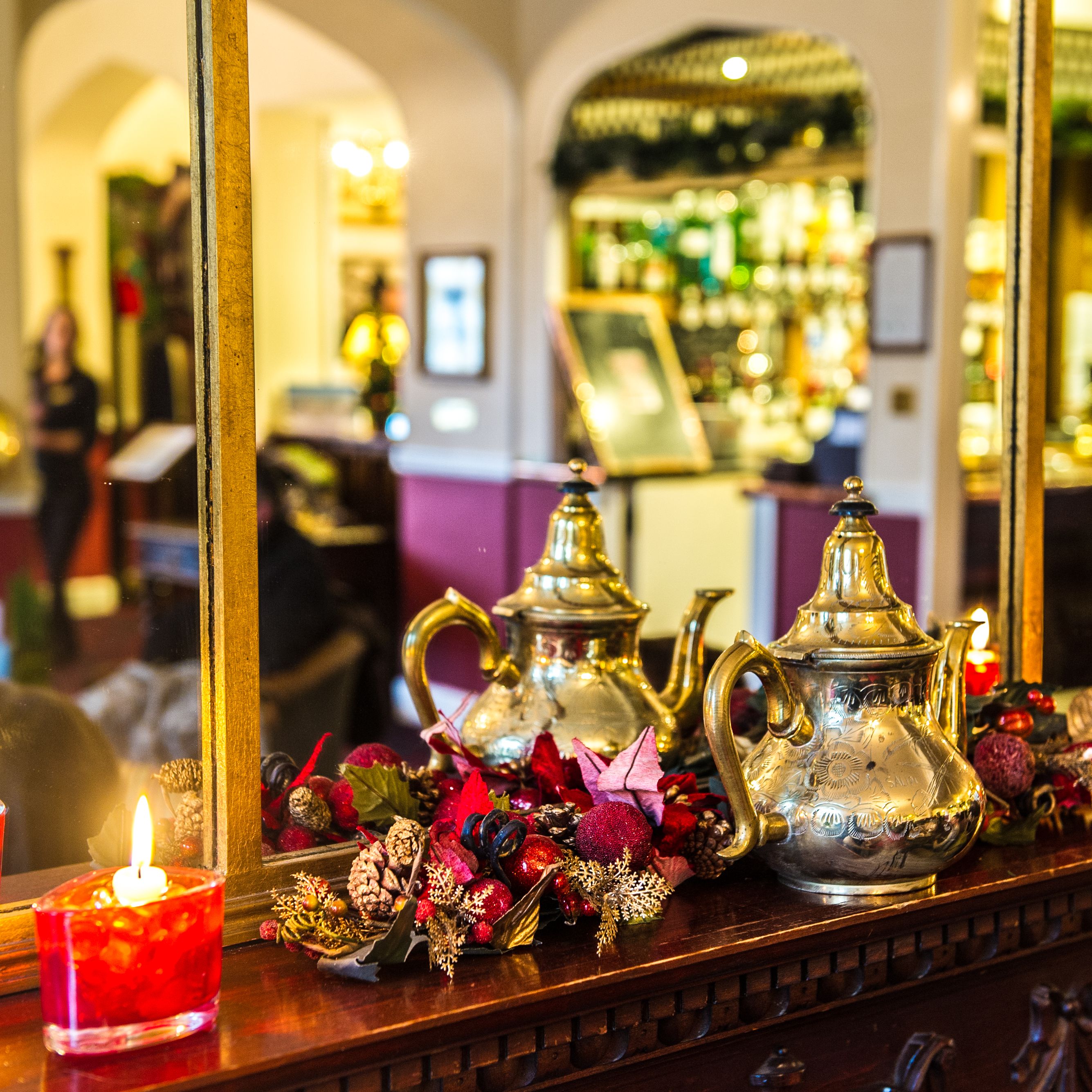 Decorative teapots and red candles on a wooden mantel with a holiday garland in front of a mirror in a warmly lit room.