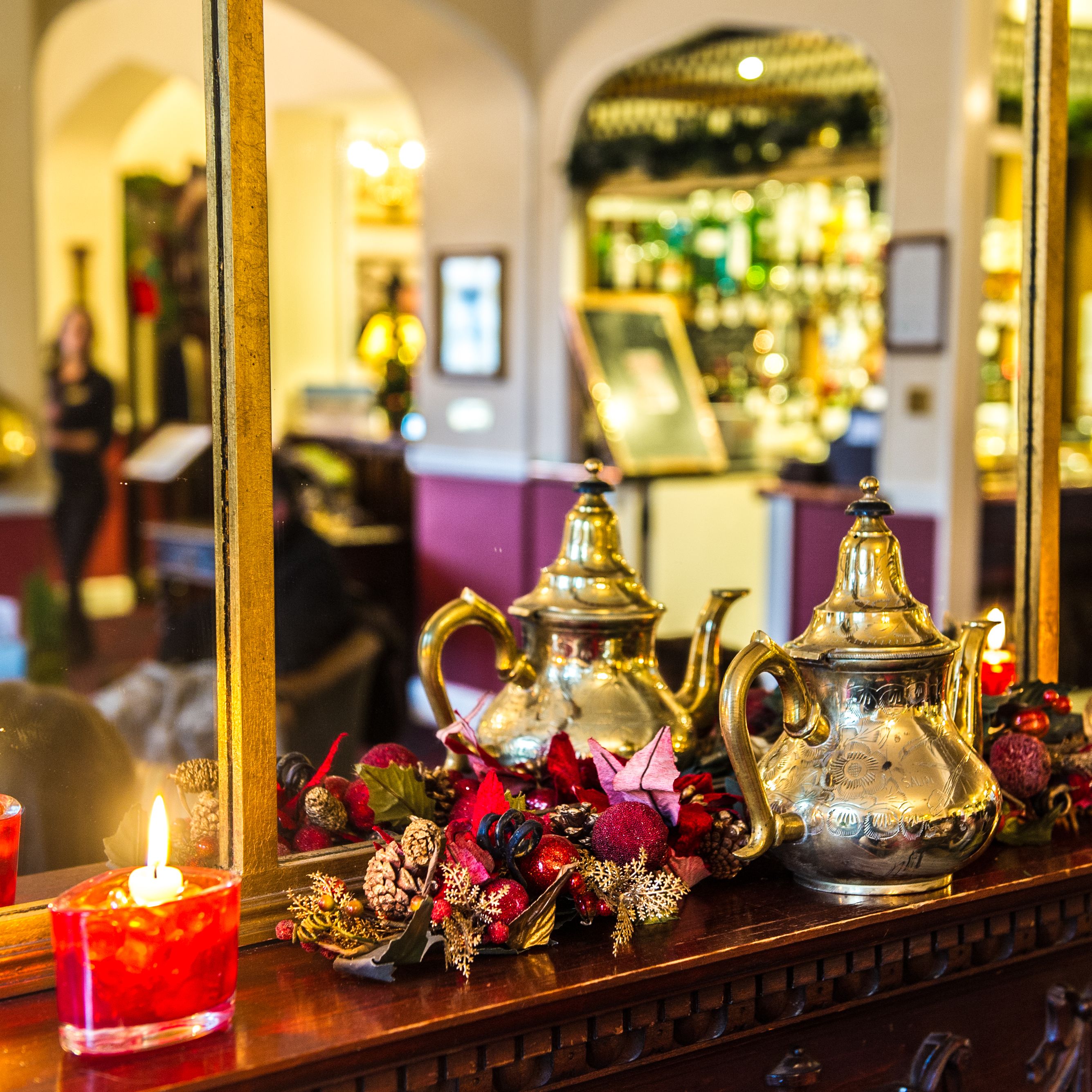Decorative teapots and red candles on a wooden mantel with a holiday garland in front of a mirror in a warmly lit room.