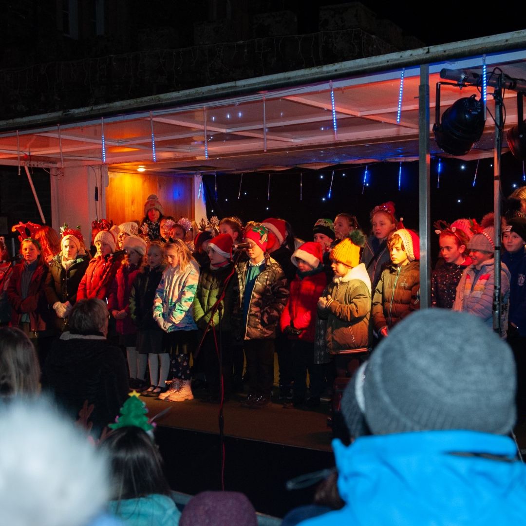Children wearing festive hats and coats singing on a stage at a nighttime outdoor event, watched by a crowd.