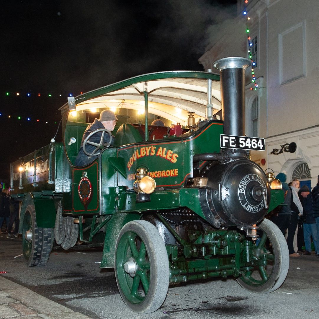 Vintage green steam engine truck labeled 'Soulby's Ales' driving through a street at night during a festival, surrounded by people.