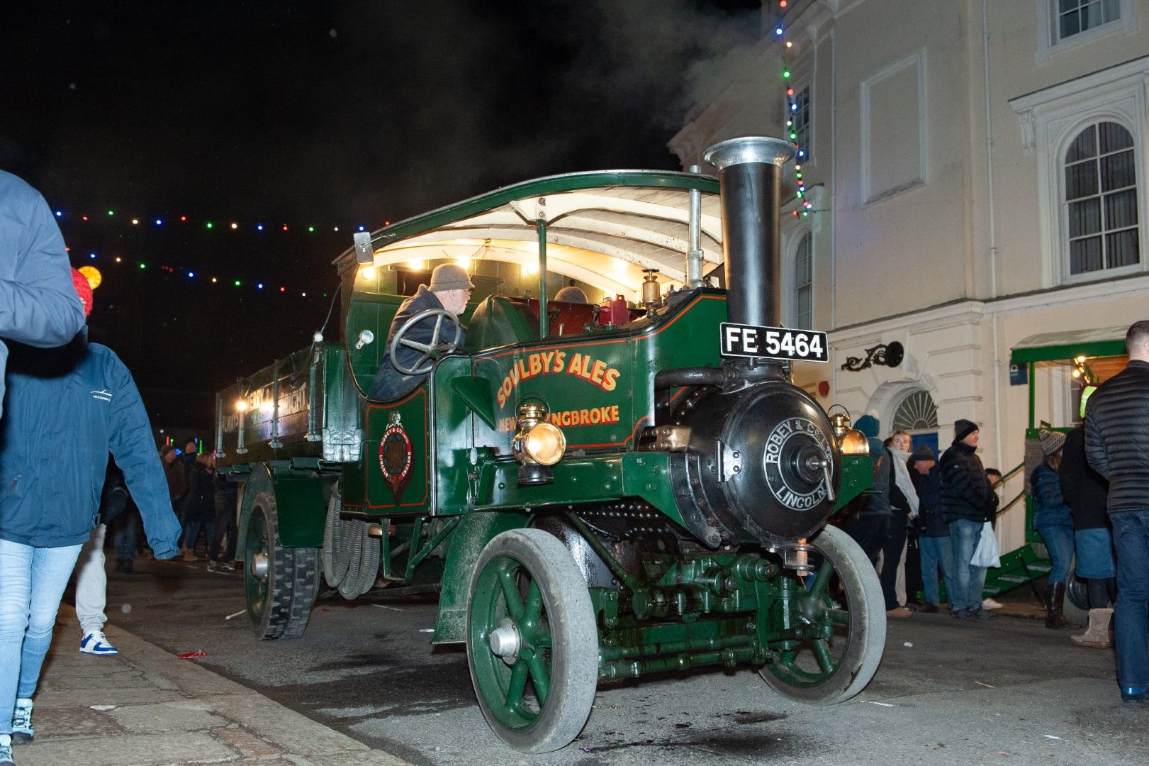 Vintage green steam engine truck labeled 'Soulby's Ales' driving through a street at night during a festival, surrounded by people.