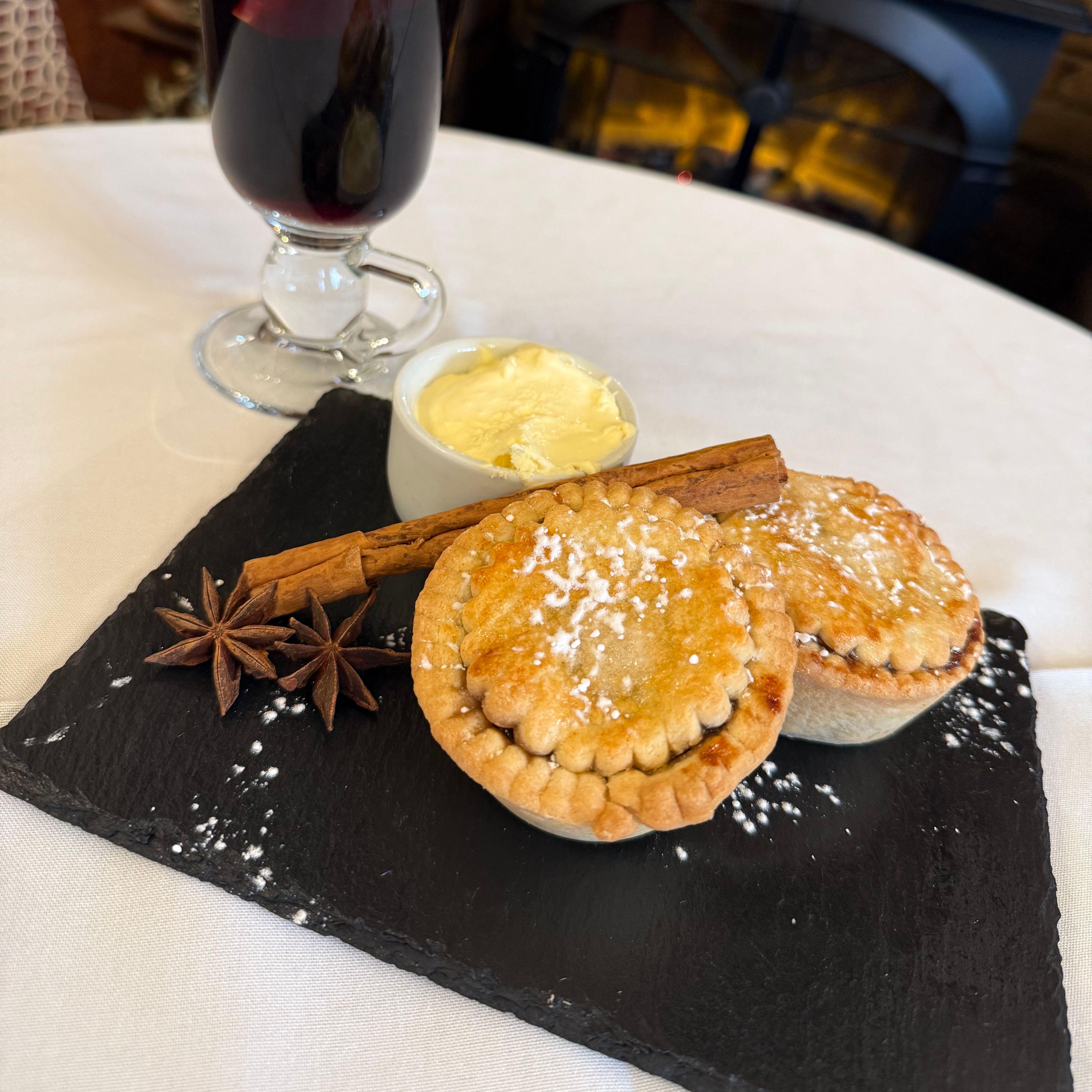 A festive serving of two mince pies with cream, cinnamon sticks, star anise, and a glass of mulled wine garnished with citrus slices.