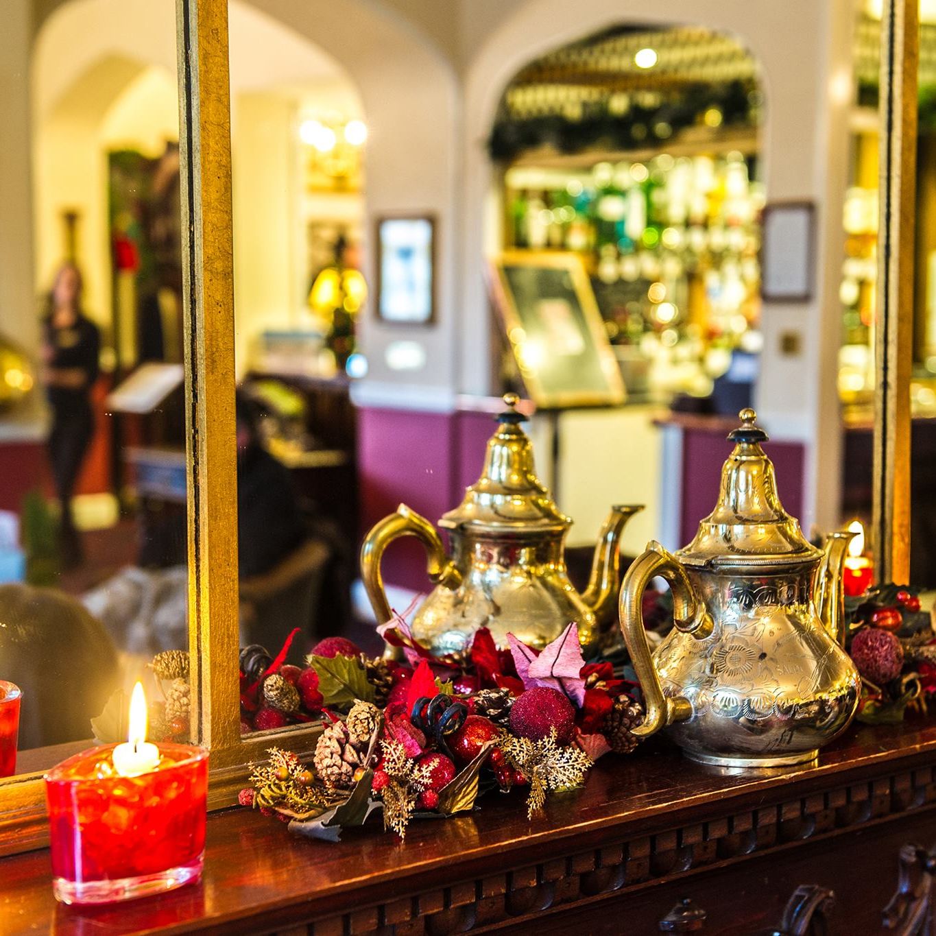 Decorative teapots, red candles, and festive garland on a wooden mantel in front of a mirror at a bar or lounge.