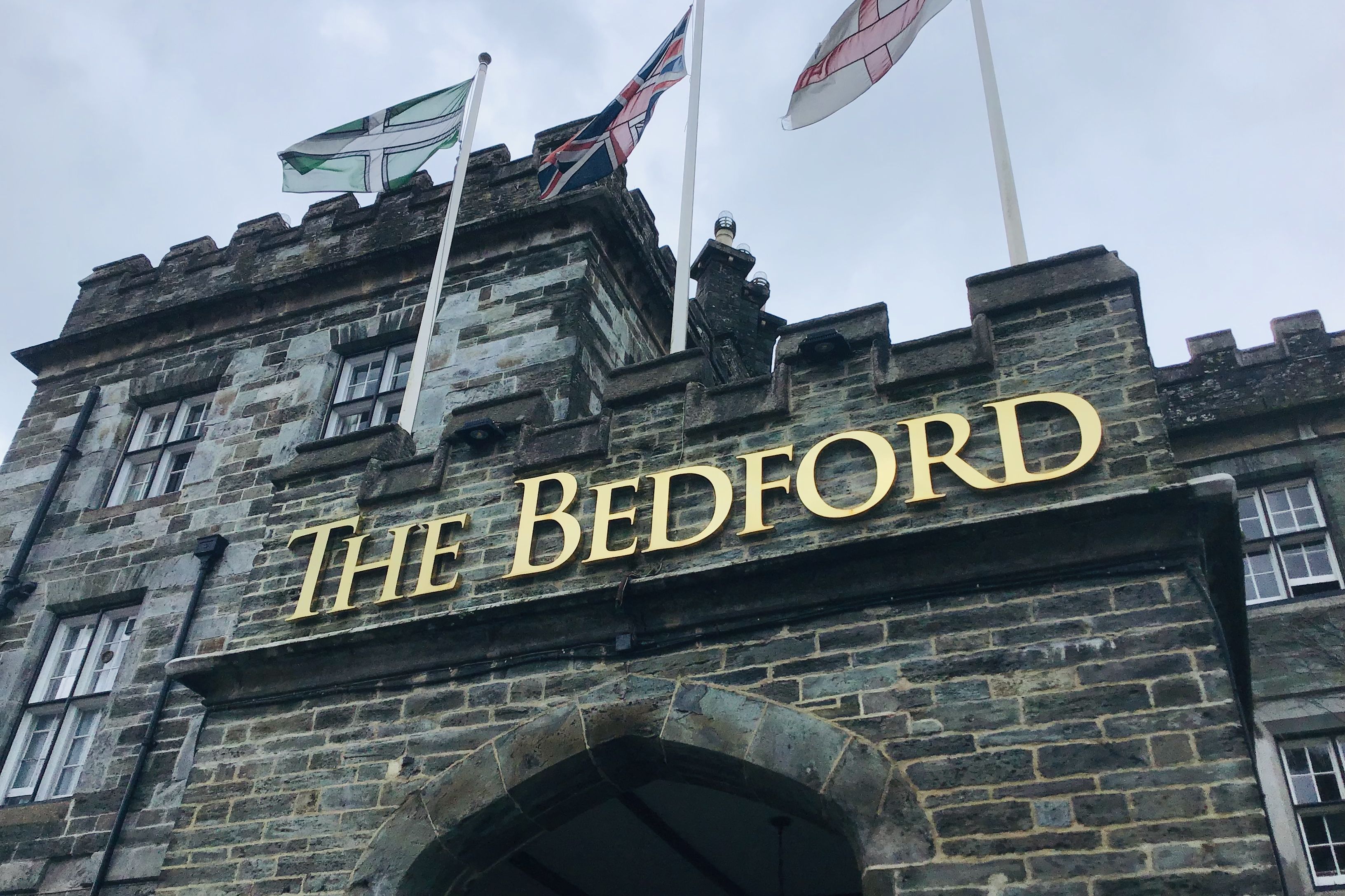 Stone building with arched entryway and gold letters spelling 'The Bedford', with three flags flying above.