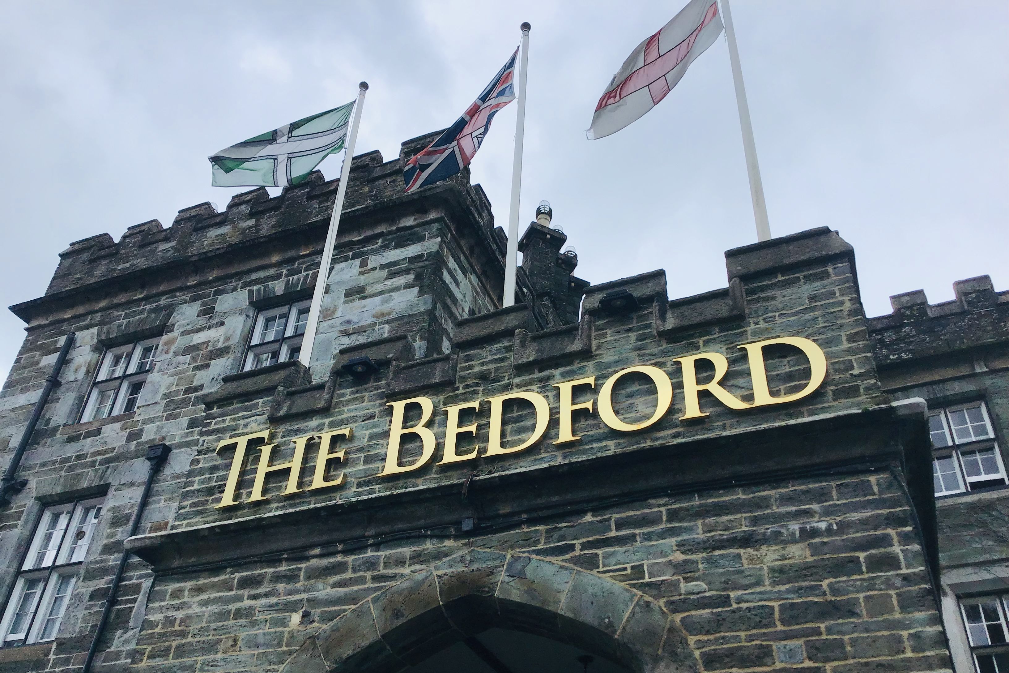 Stone building with arched entryway and gold letters spelling 'The Bedford', with three flags flying above.
