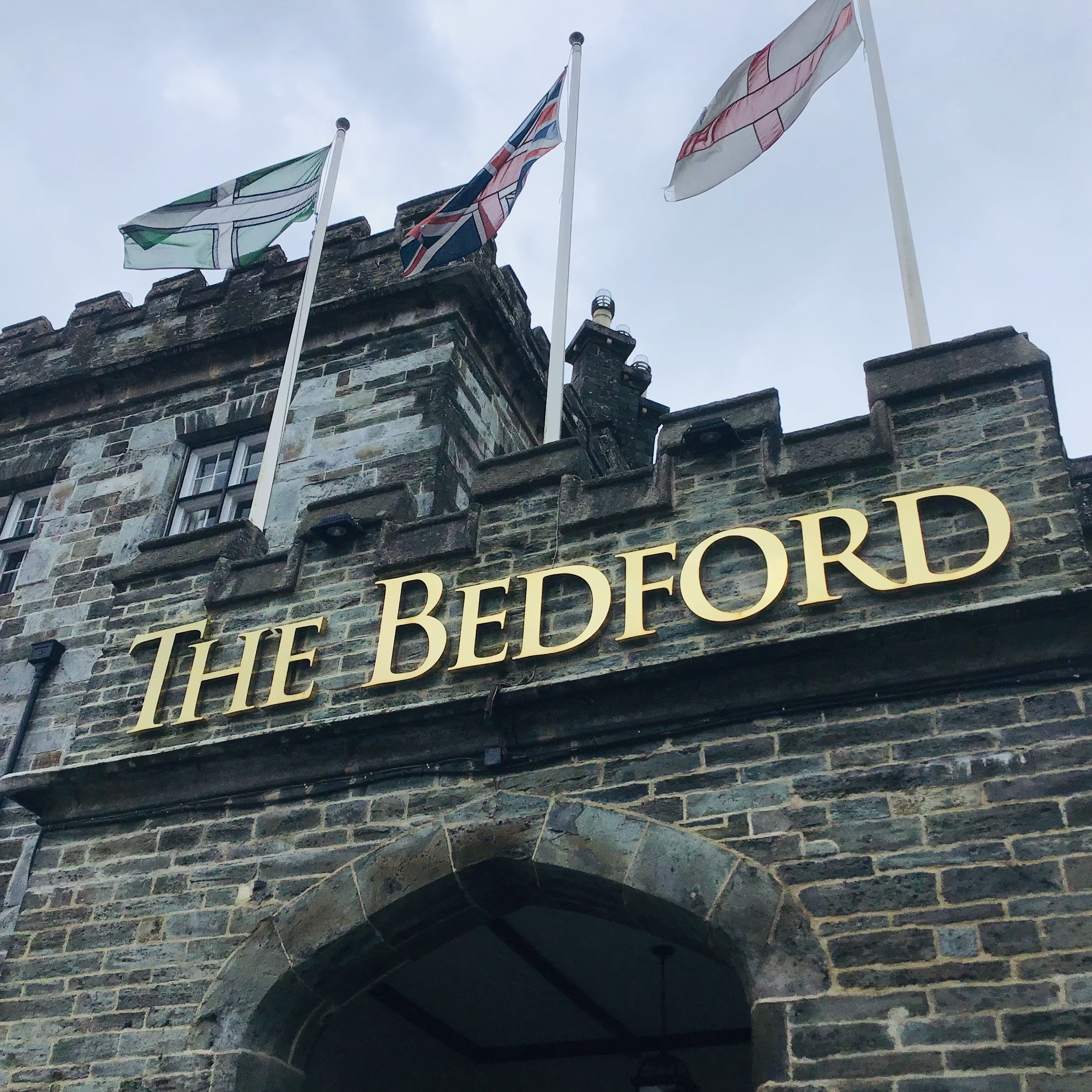 Stone building with arched entryway and gold letters spelling 'The Bedford', with three flags flying above.