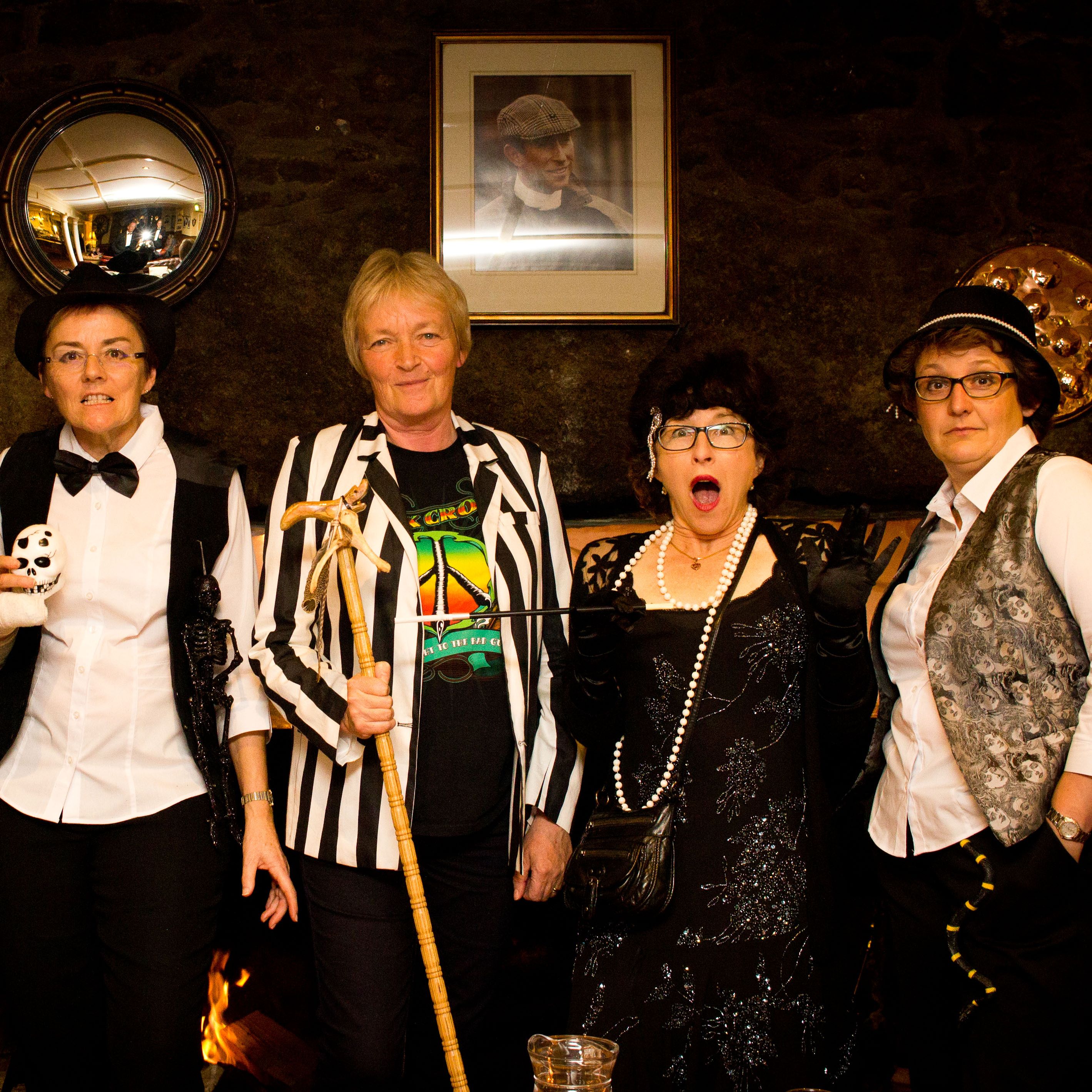 Group of six adults dressed in various murder mystery themed costumes, standing together indoors.