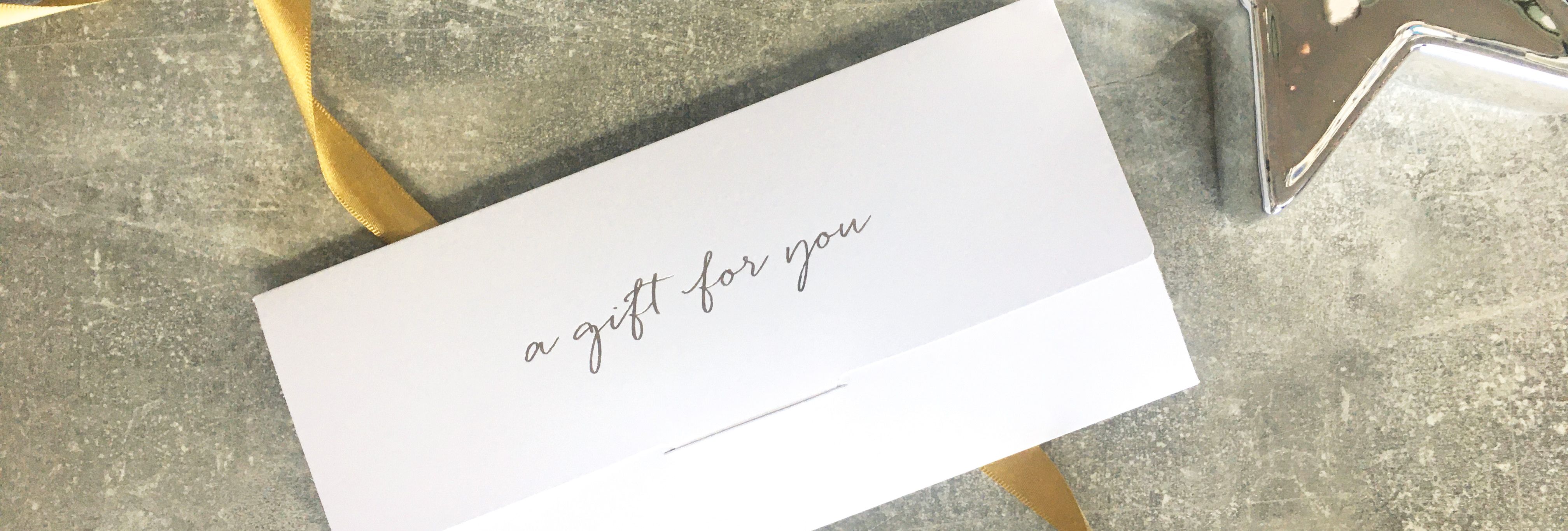 White gift voucher envelope with 'a gift for you' text, next to a yellow ribbon and a shiny silver star on a textured surface.