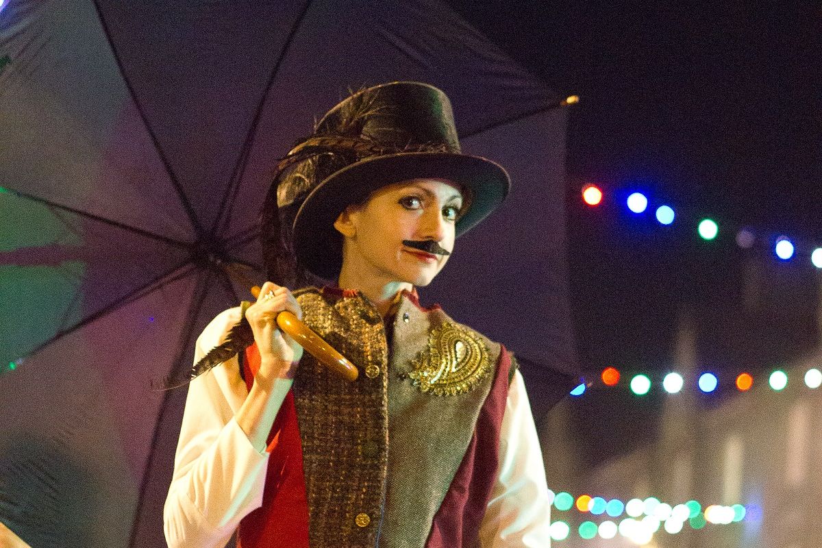 Person in Victorian costume with a mustache and top hat holding an umbrella at night, with colorful string lights in the background.