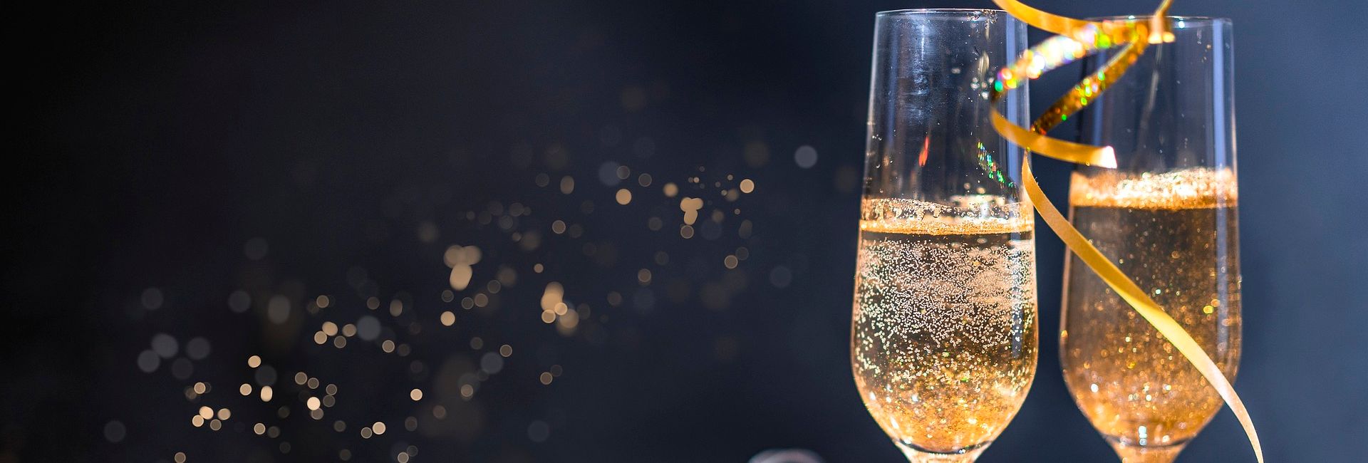 Two champagne glasses with golden ribbon, bottle of champagne, and festive lights