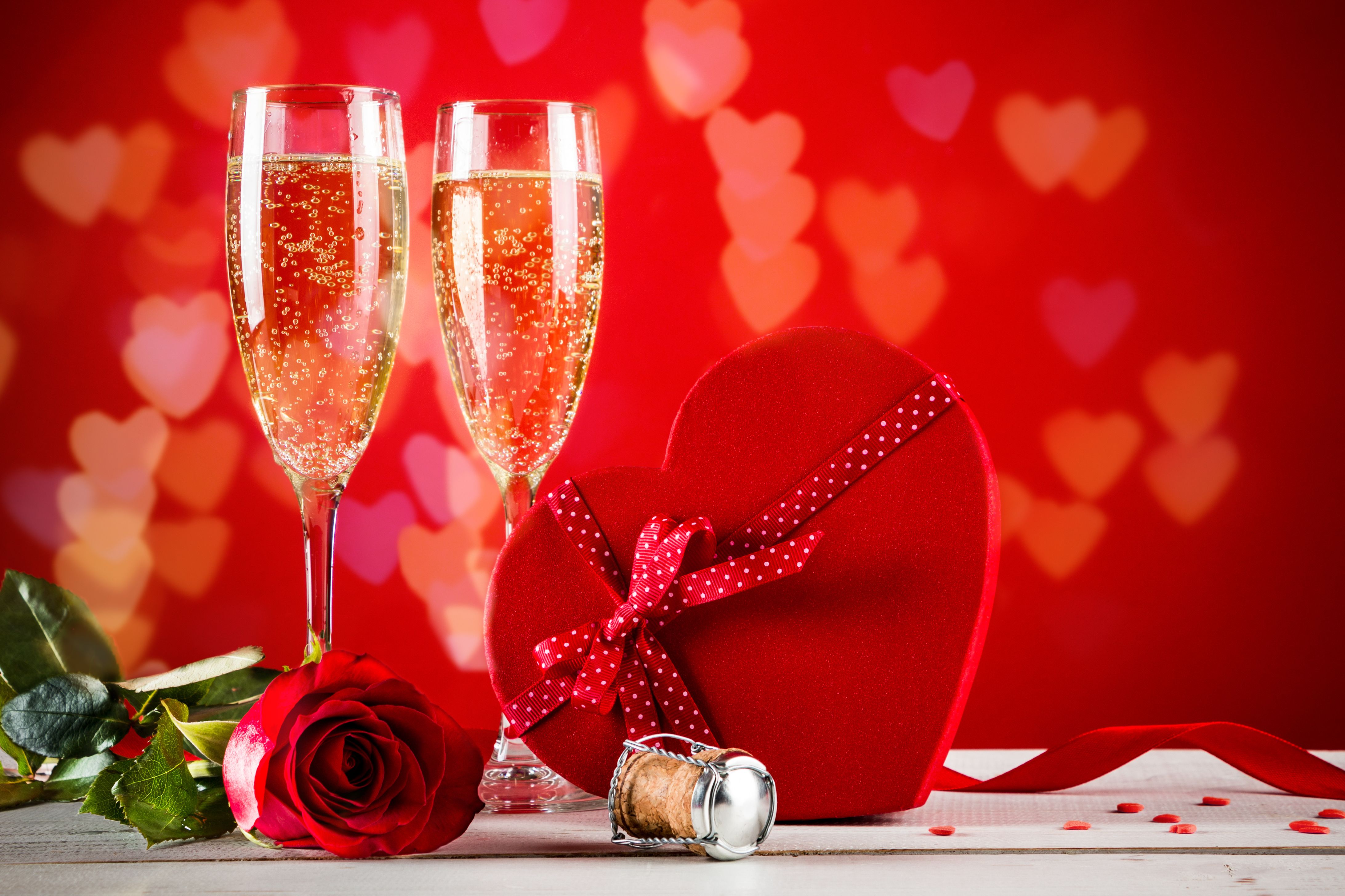 Romantic setting with two champagne glasses, a red rose, and a heart-shaped gift box on a red background with heart-shaped bokeh.