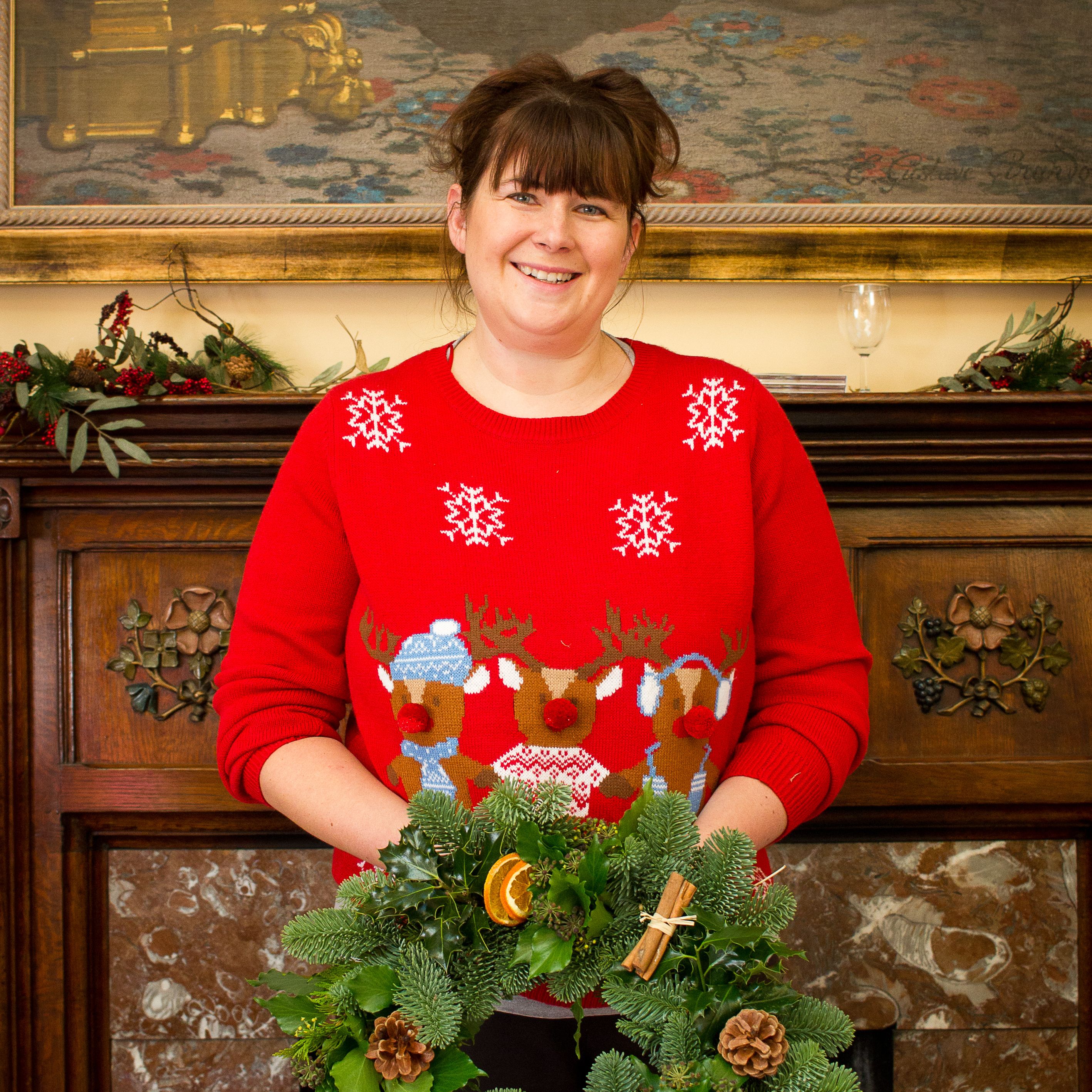 Woman in a festive red sweater standing behind a table making a holiday wreath decorated with pine, orange slices, cinnamon sticks, and pinecones.