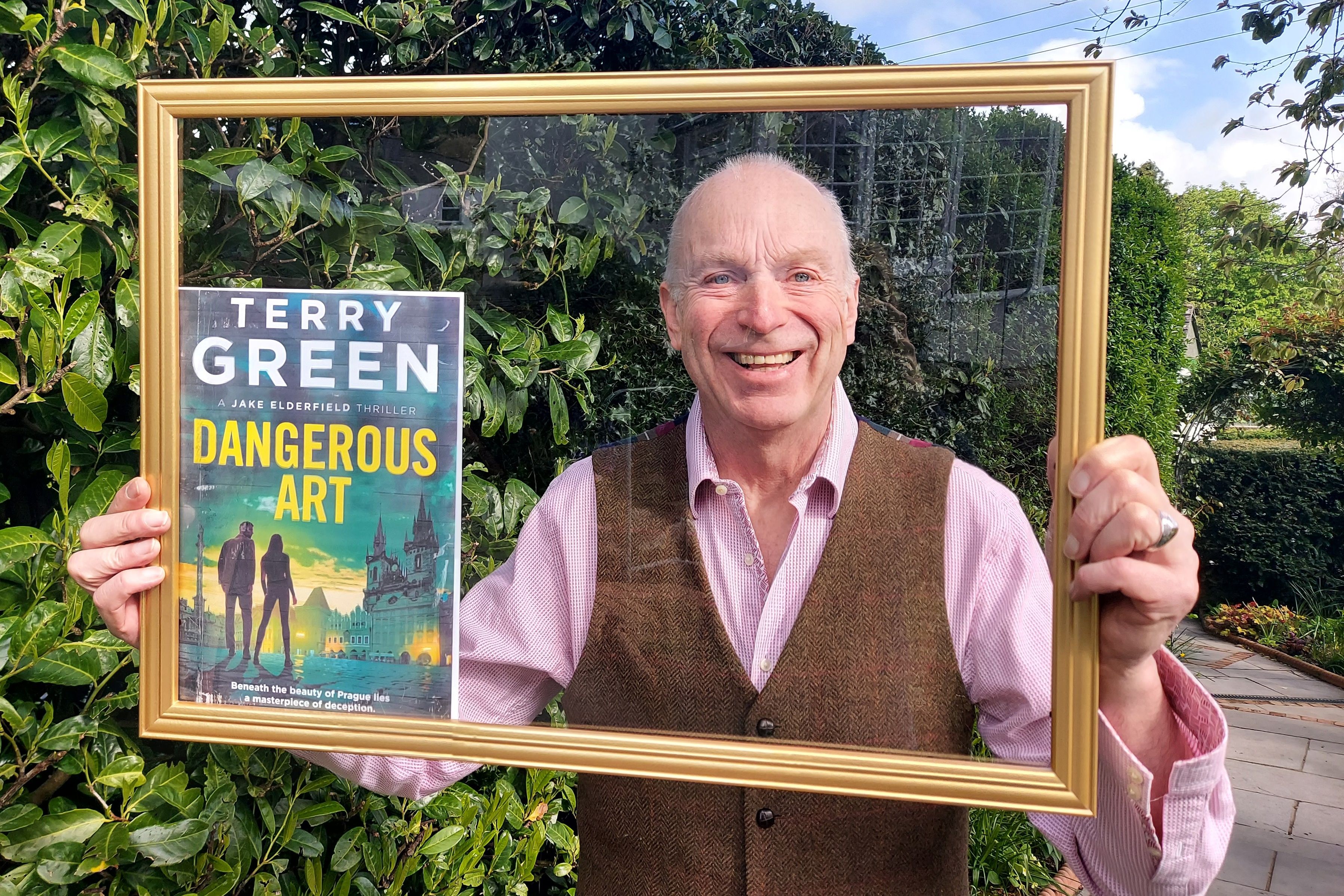 Smiling man holding a golden picture frame and a book titled 'Dangerous Art' by Terry Green in a garden.