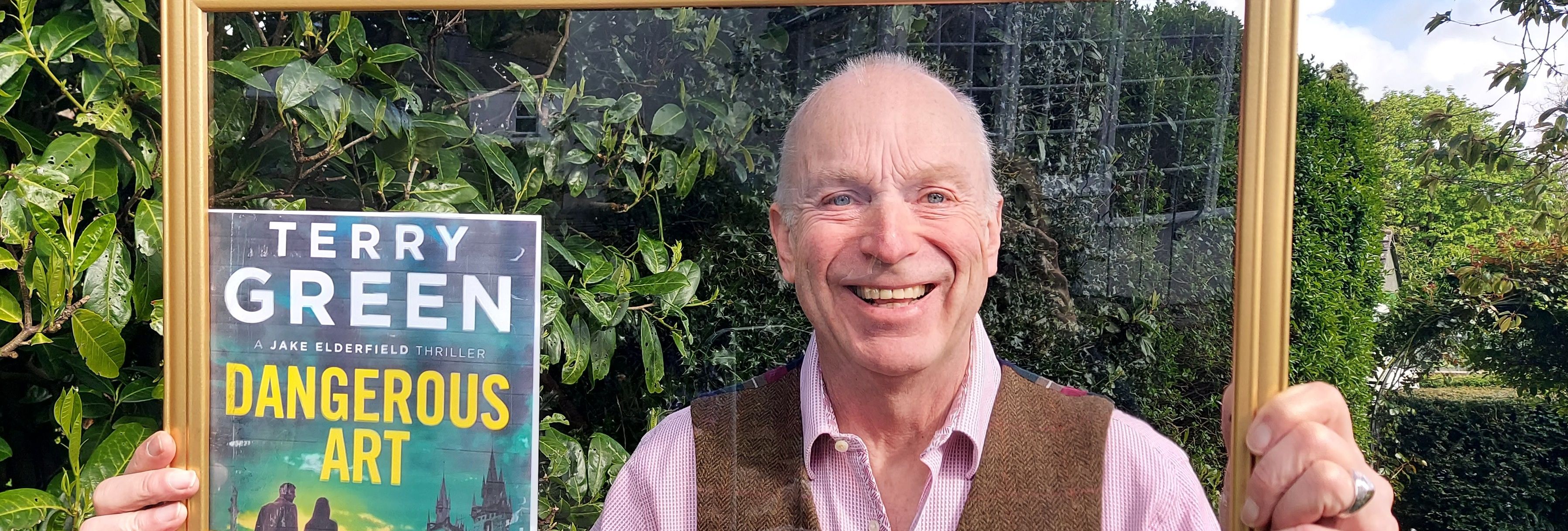 Smiling man holding a golden picture frame and a book titled 'Dangerous Art' by Terry Green in a garden.