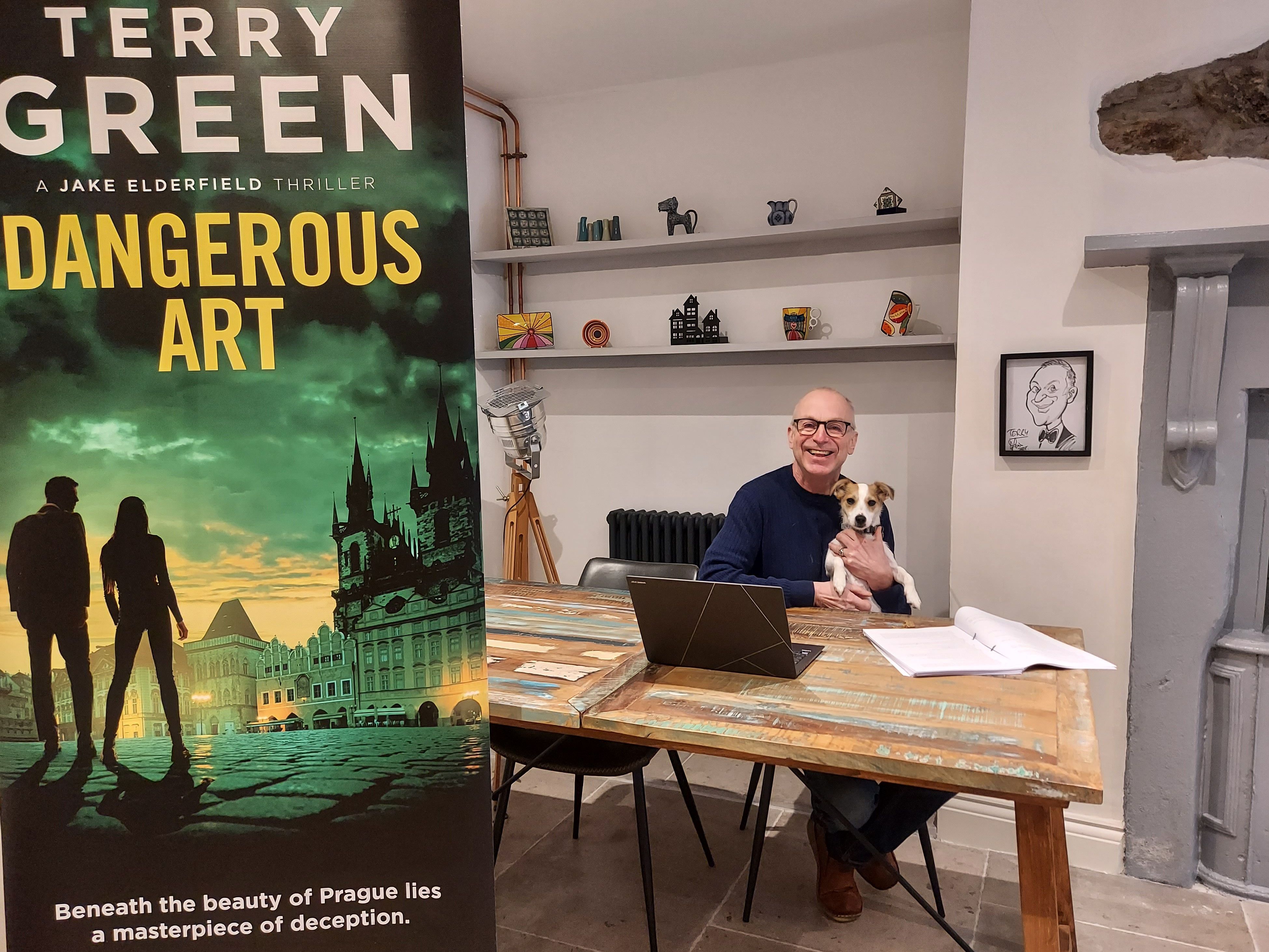 A man sitting at a rustic wooden desk with a small dog on his lap, smiling at the camera. To the left is a large banner for a thriller novel called 'Dangerous Art' by Terry Green.