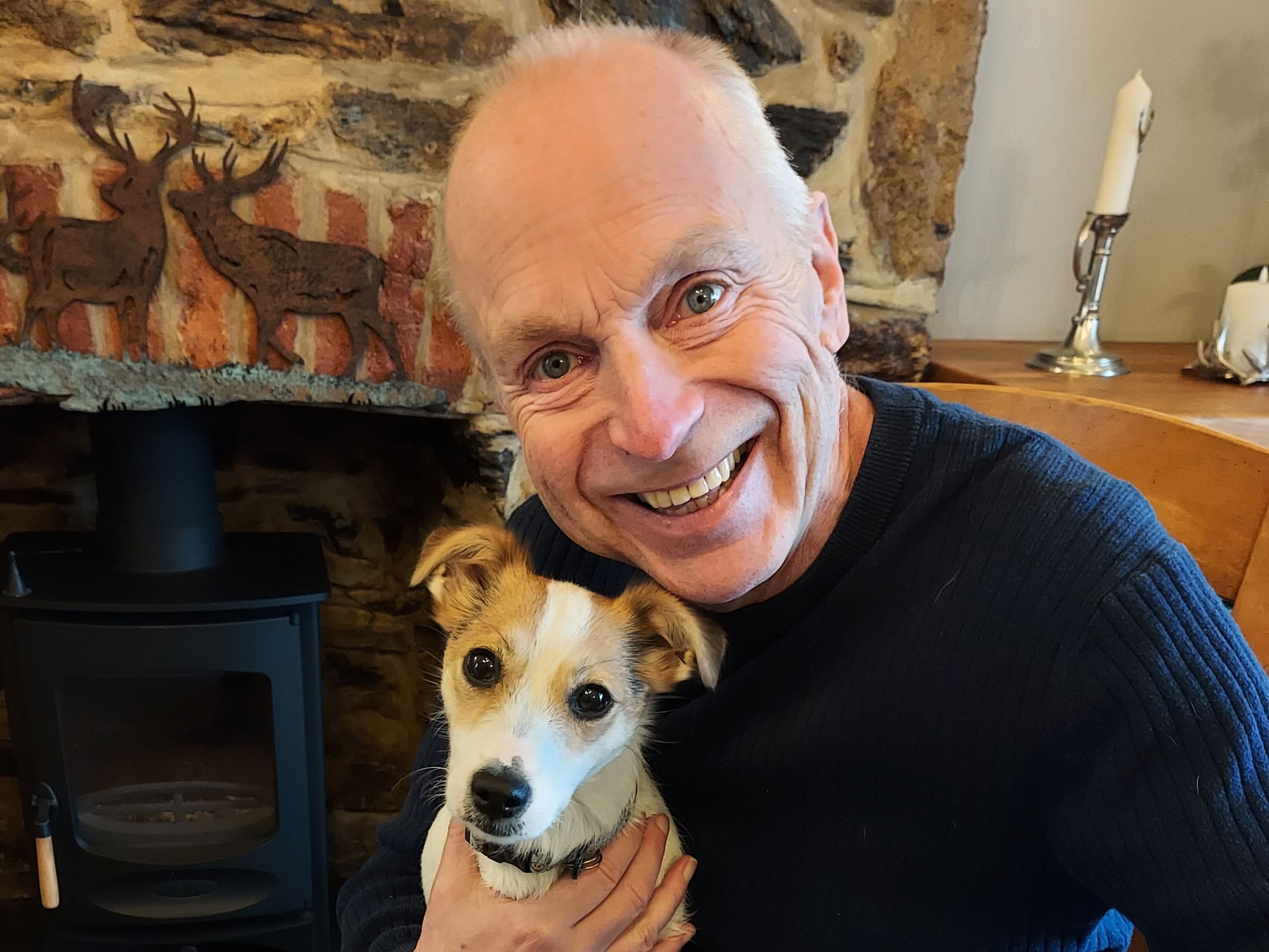 Smiling elderly man holding a small dog in a cozy room with a stone fireplace in the background.