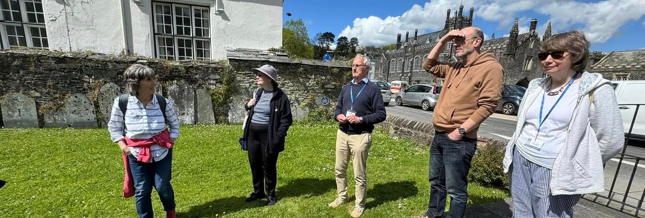 A group of five adults standing and talking outside near a stone building with large windows on a sunny day.