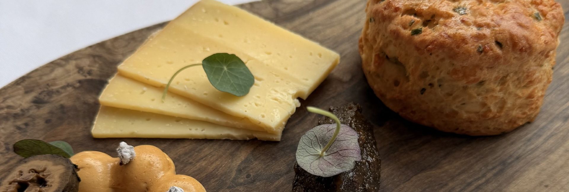 Wooden serving board with a scone, cheese slices, a mousse ring, and a dark spread garnished with herbs