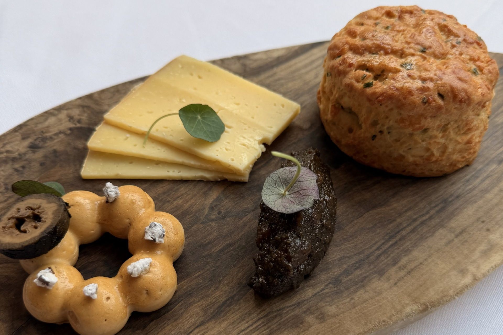 Wooden serving board with a scone, cheese slices, a mousse ring, and a dark spread garnished with herbs
