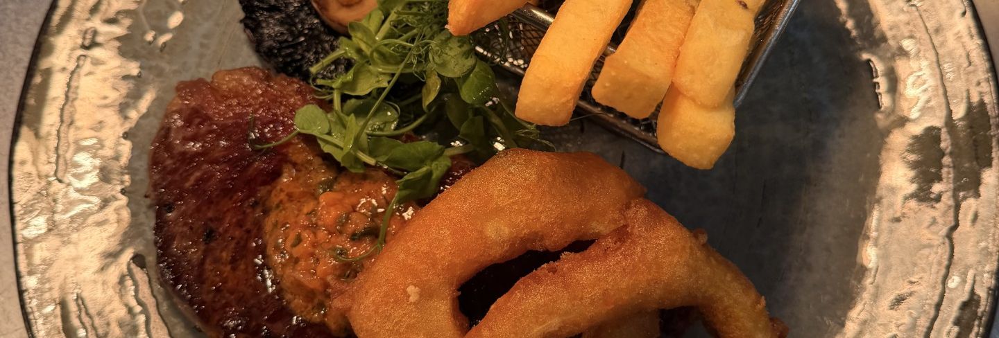 Plate of steak meal with thick-cut fries, onion rings, grilled mushroom, tomato, and greens.
