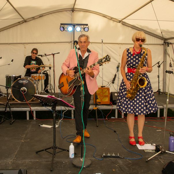 A four-member band performing on stage under a white tent. The members are playing electric guitar, drums, saxophone, and bass guitar.