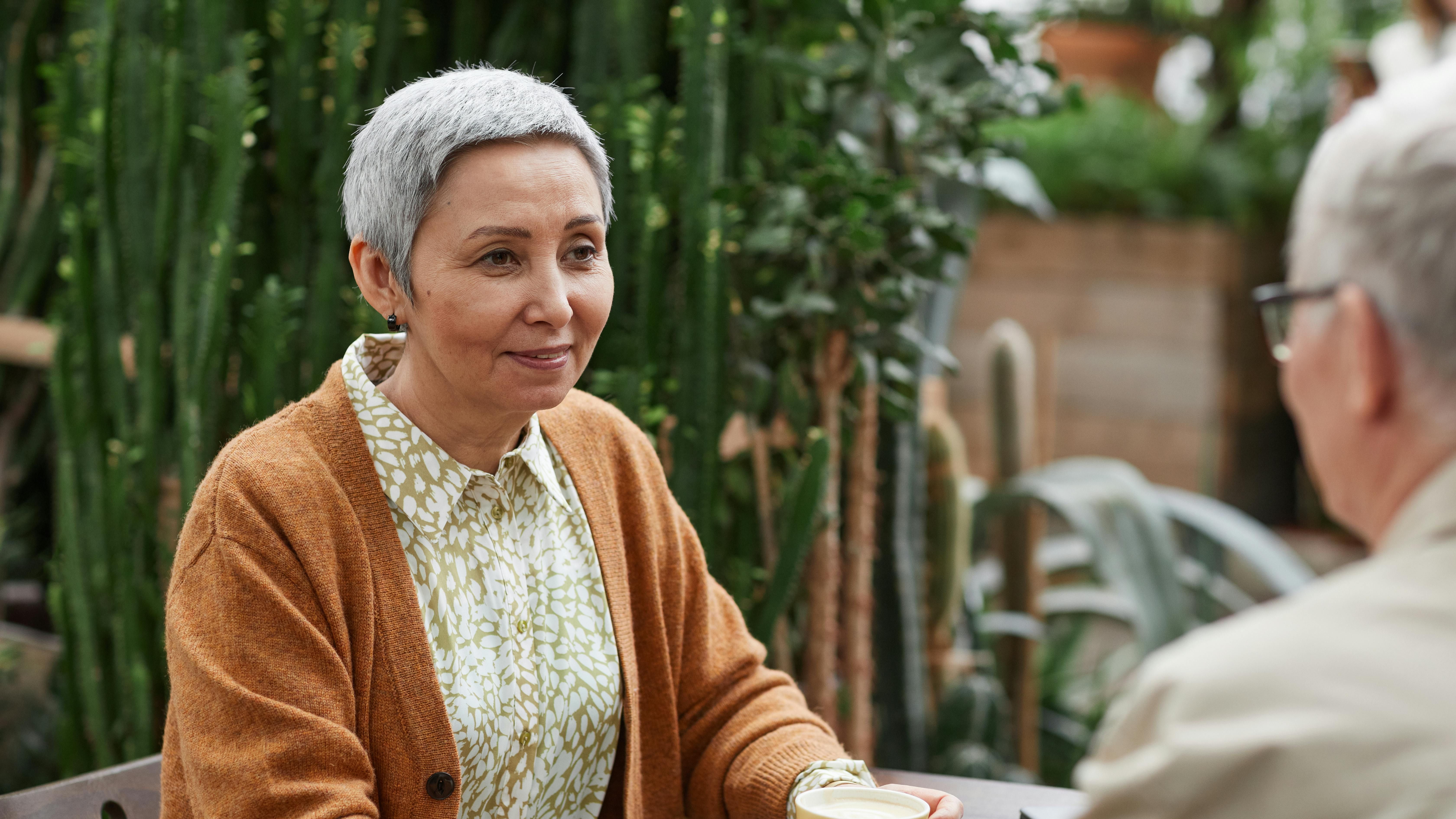 Older woman with short gray hair smiles while holding a cup of coffee across from a man in a lush, green, plant-filled café.