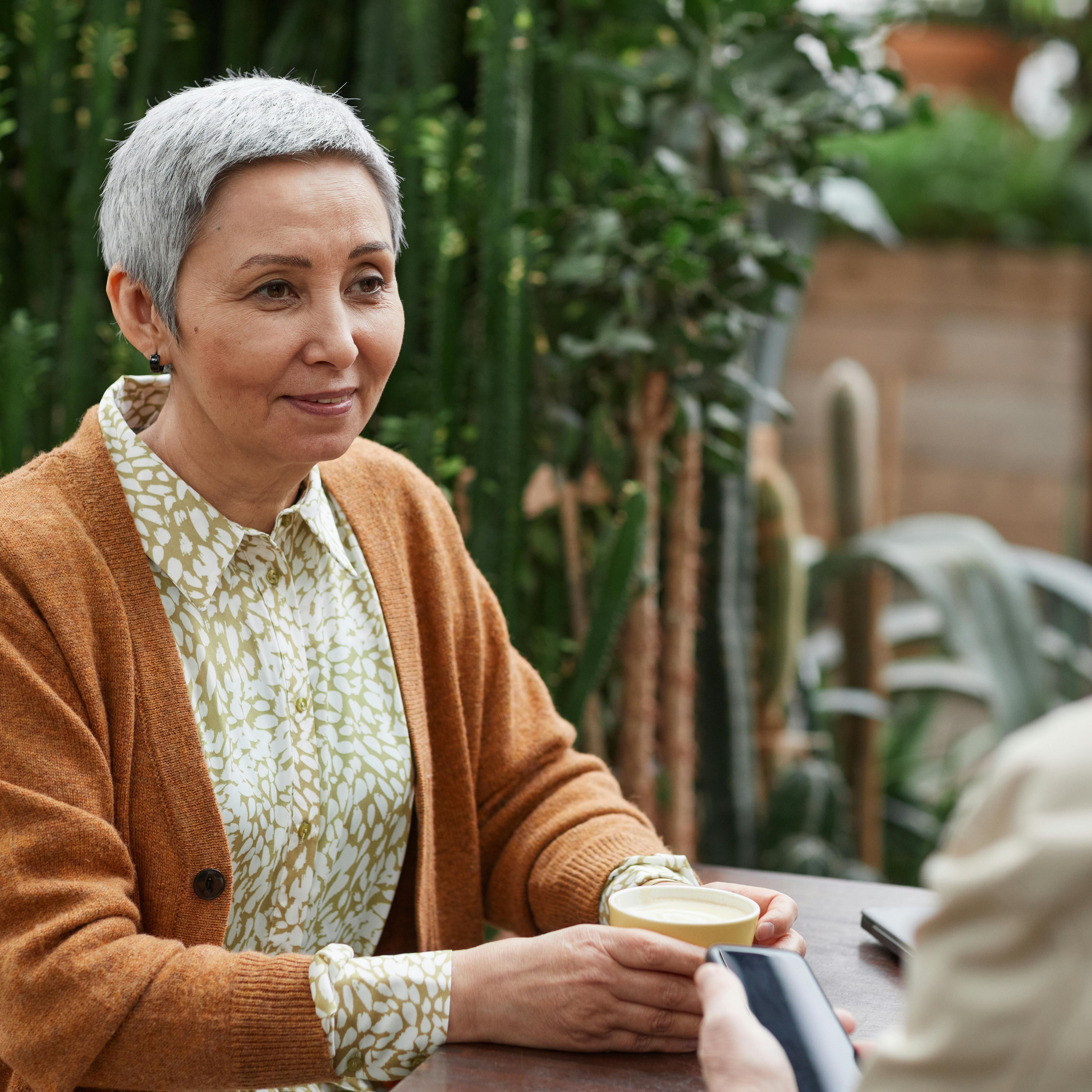 Older woman with short gray hair smiles while holding a cup of coffee across from a man in a lush, green, plant-filled café.