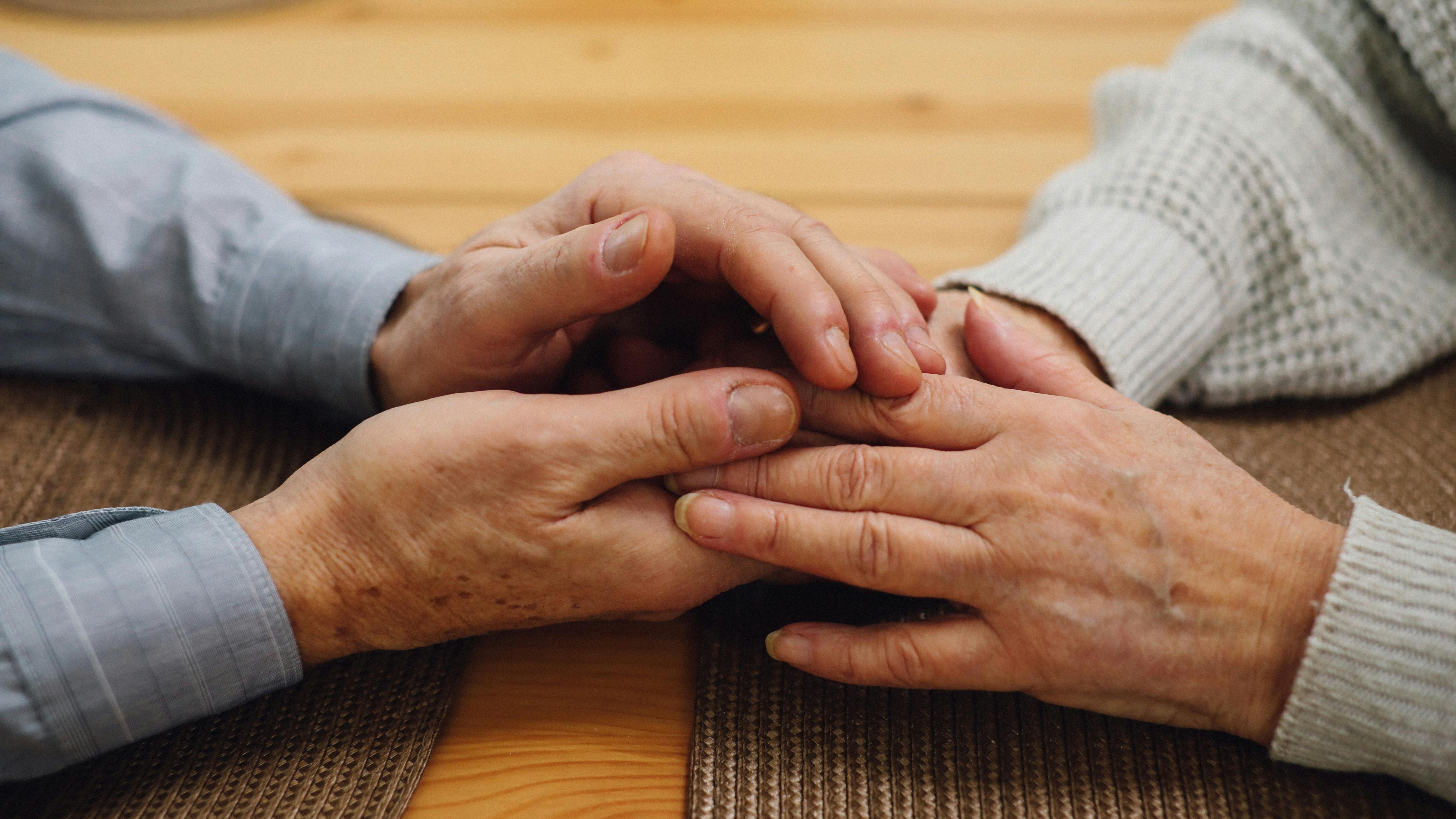 Close-up of two elderly people holding hands across a wooden table, conveying comfort, care, and emotional connection in a warm, intimate moment.
