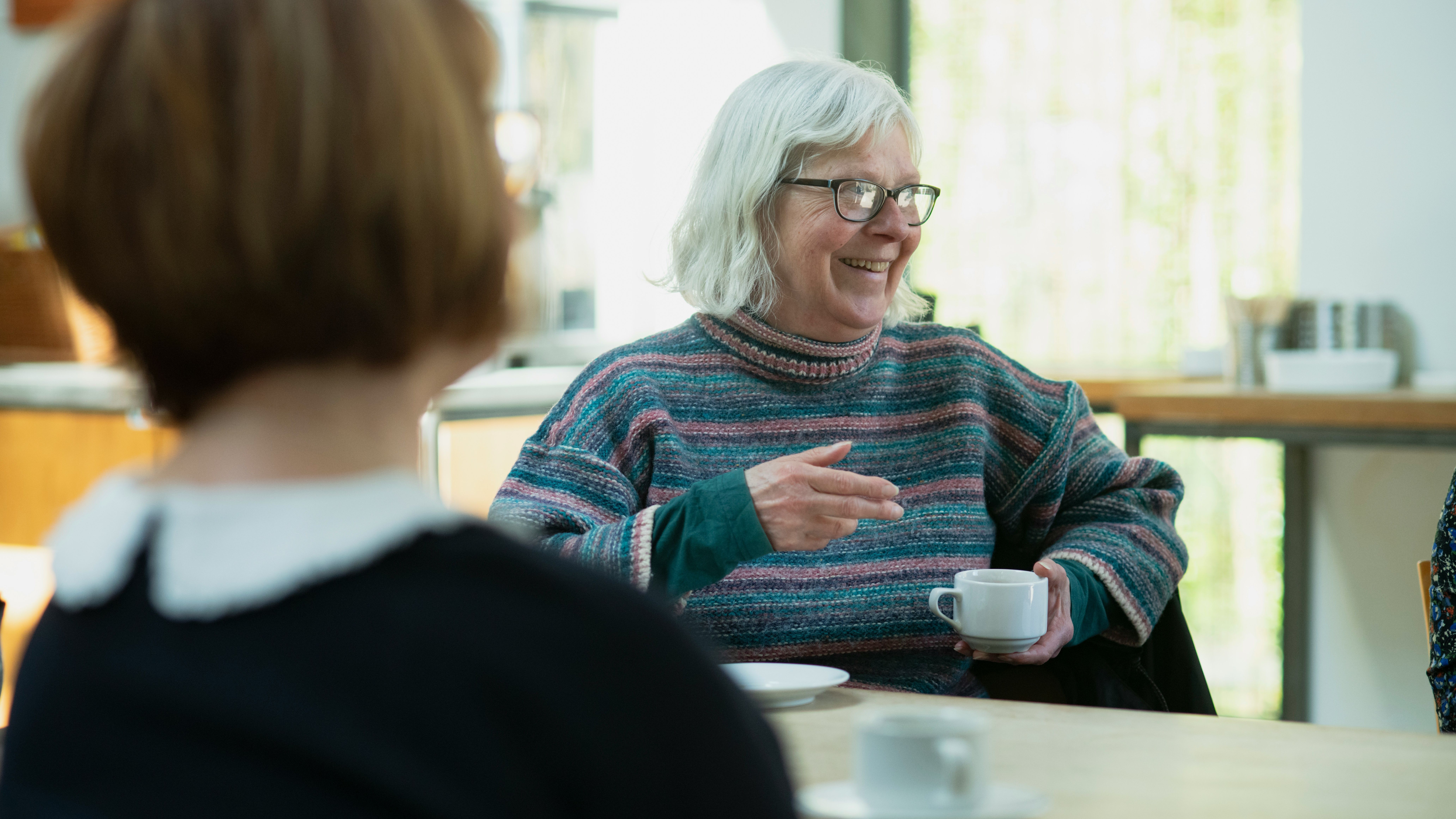 Smiling elderly woman with white hair and glasses enjoying a conversation over coffee in a cosy café setting.