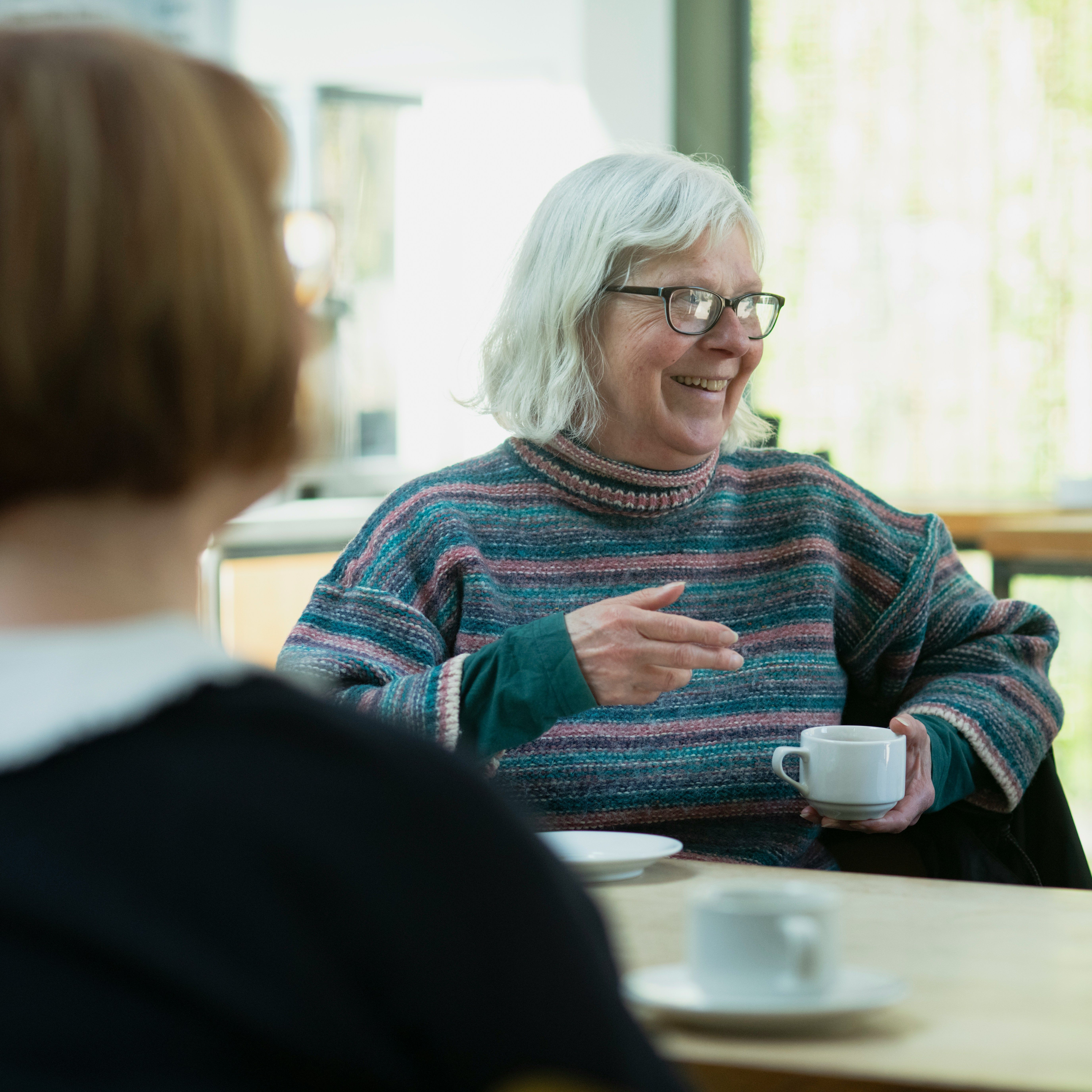 Smiling elderly woman with white hair and glasses enjoying a conversation over coffee in a cosy café setting.