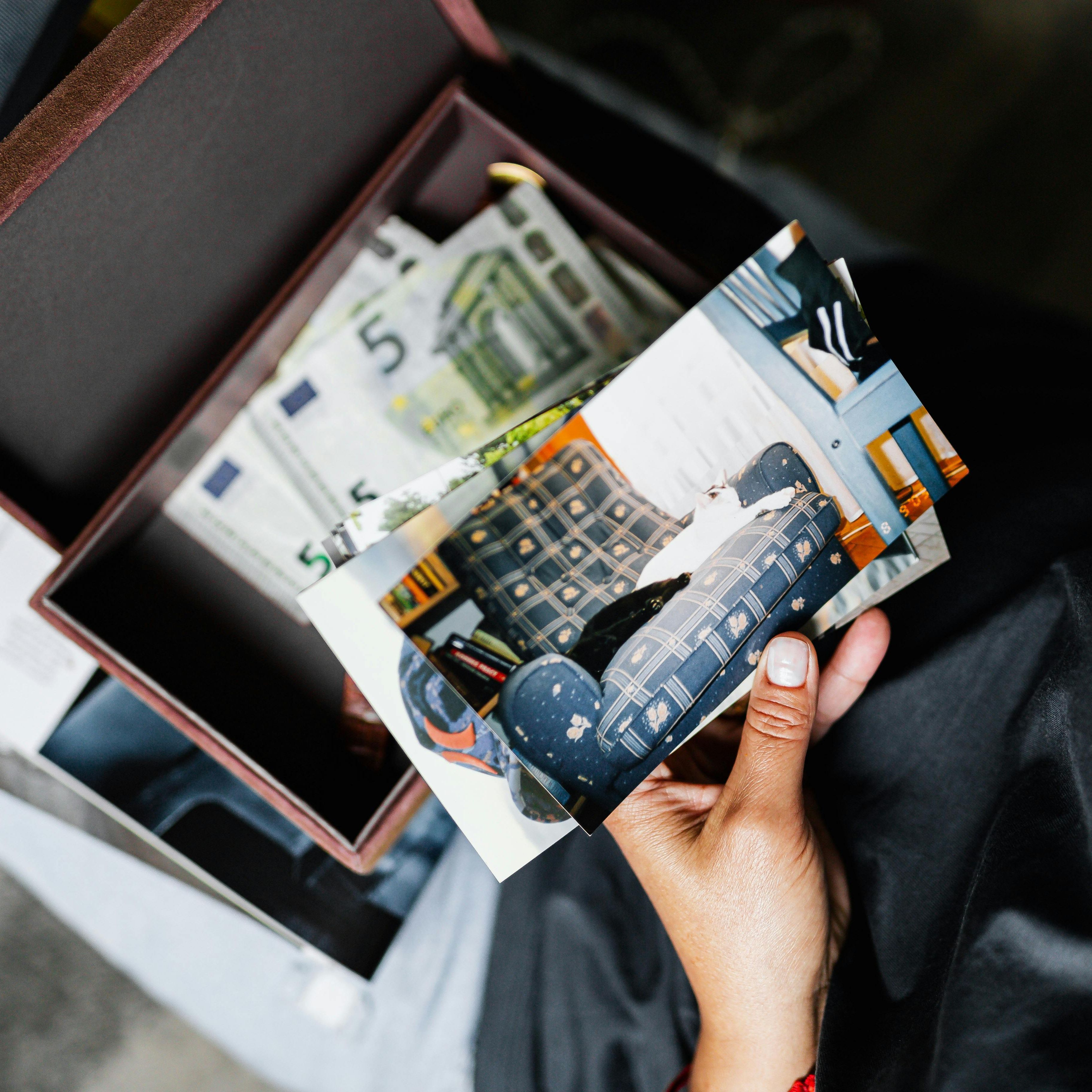 A person holding old printed photos above an open box filled with euro banknotes and more photographs.