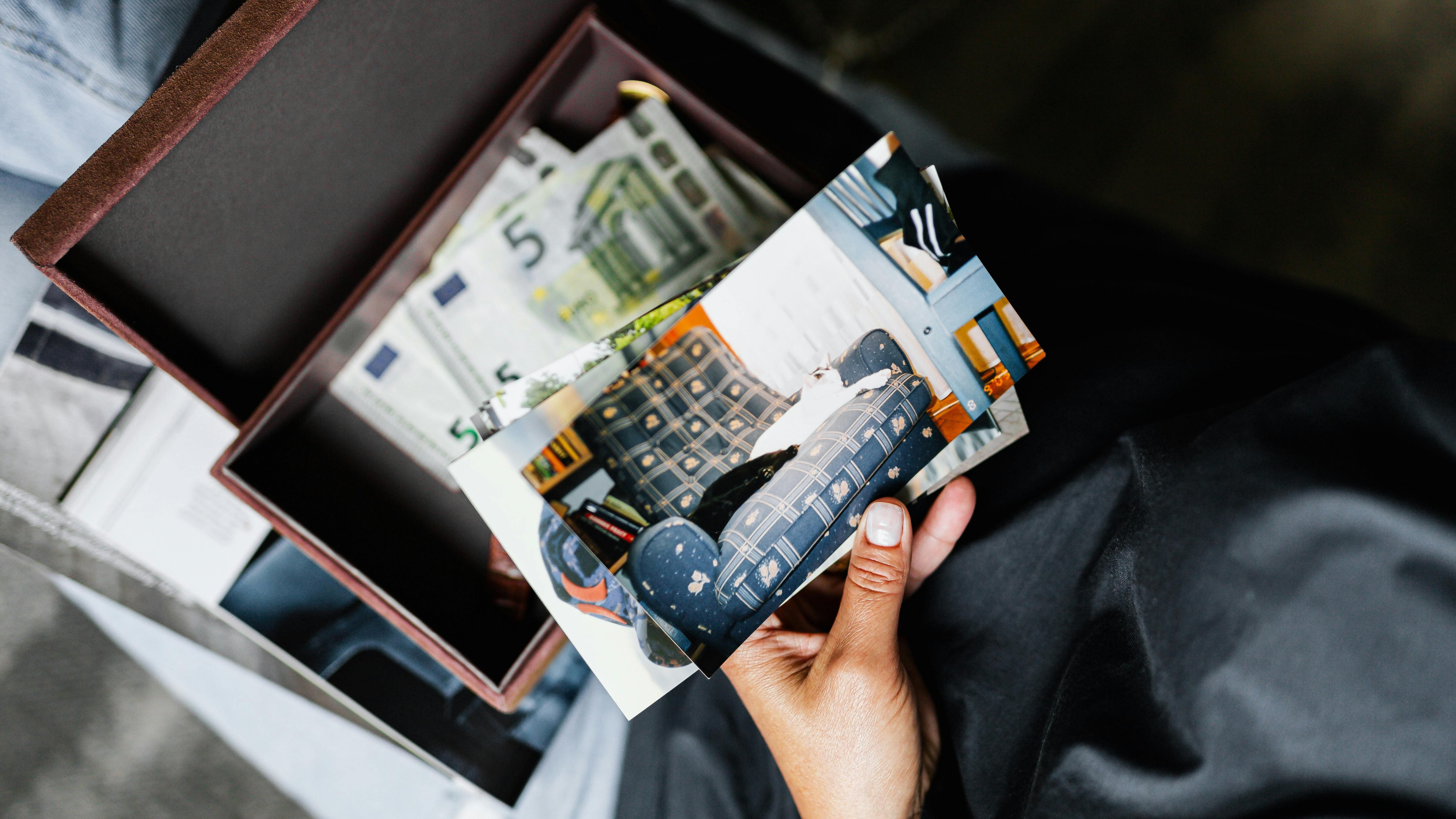 A person holding old printed photos above an open box filled with euro banknotes and more photographs.
