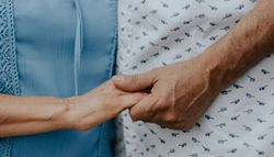 Close-up of two people holding hands, one wearing a blue blouse and the other a white shirt with a pattern of small glasses.