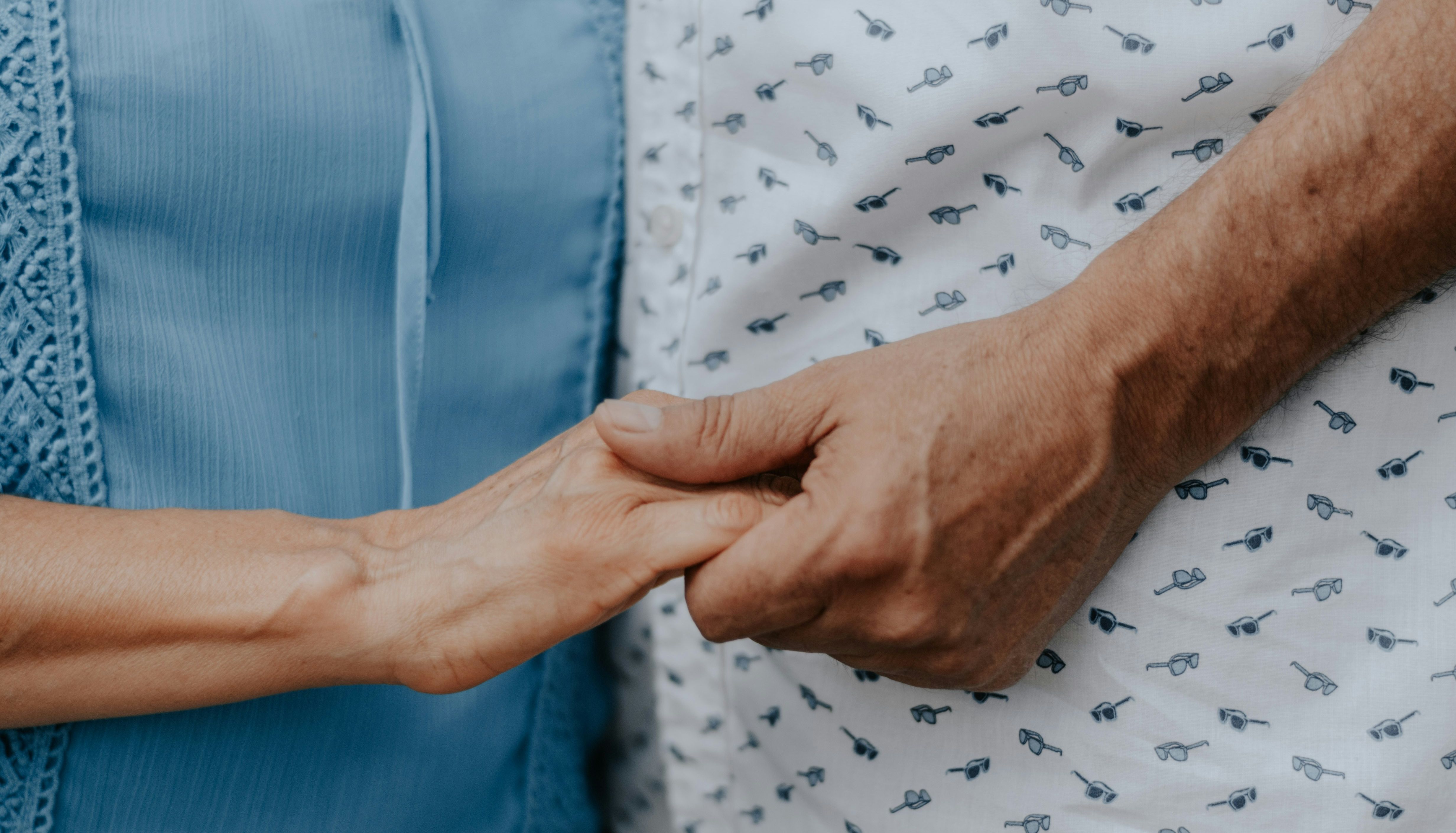 Close-up of two people holding hands, one wearing a blue blouse and the other a white shirt with a pattern of small glasses.