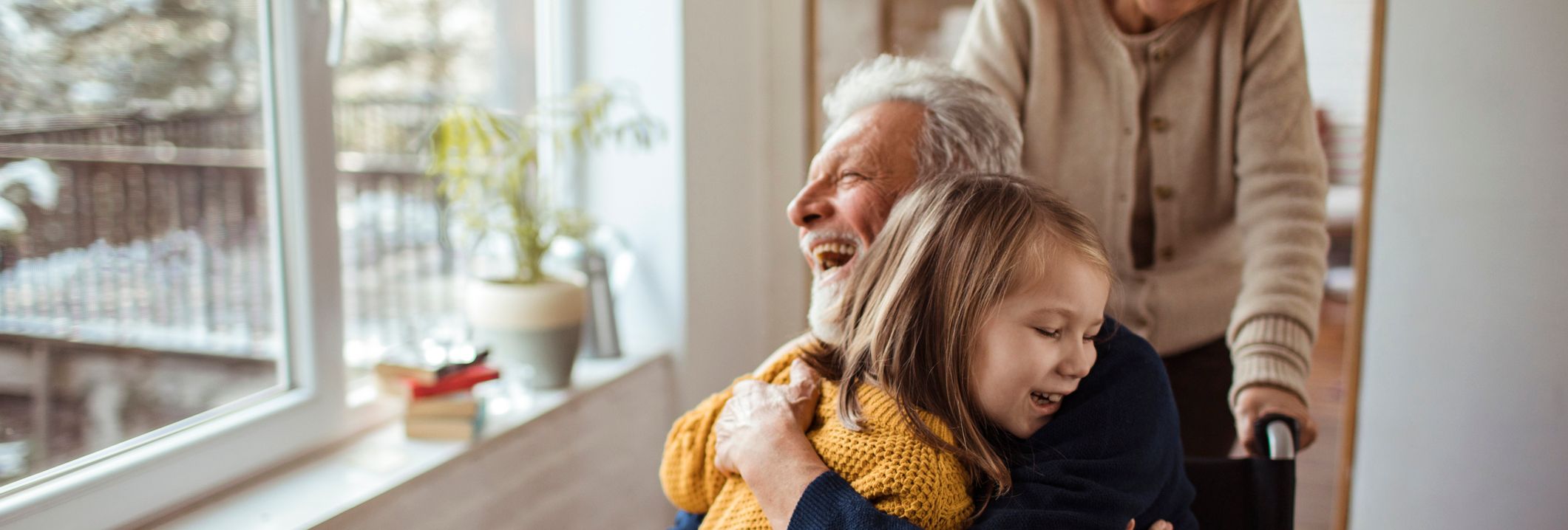 A joyful elderly man in a wheelchair hugs a laughing young girl as an older woman smiles behind them, indoors near a bright window.