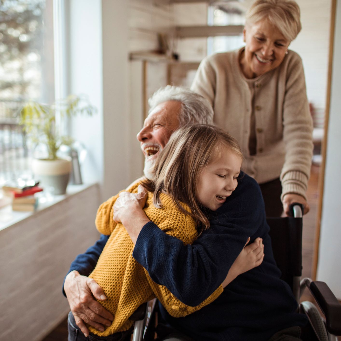 A joyful elderly man in a wheelchair hugs a laughing young girl as an older woman smiles behind them, indoors near a bright window.