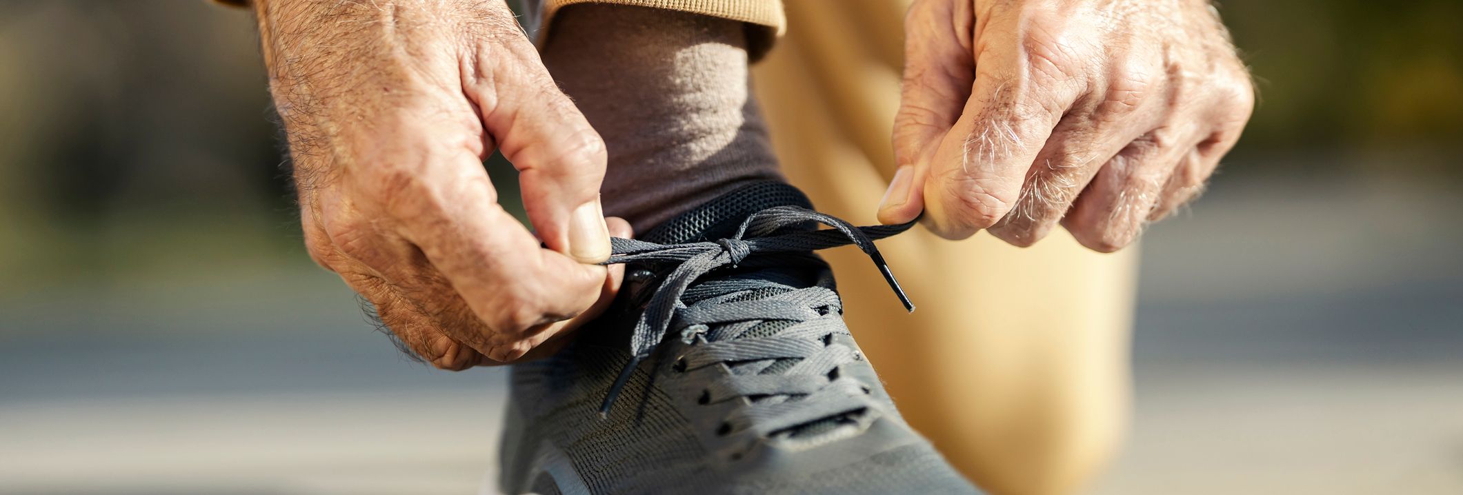 Close-up of an older adult tying the laces on a black athletic shoe while kneeling on pavement, wearing tan pants and a yellow sweatshirt.