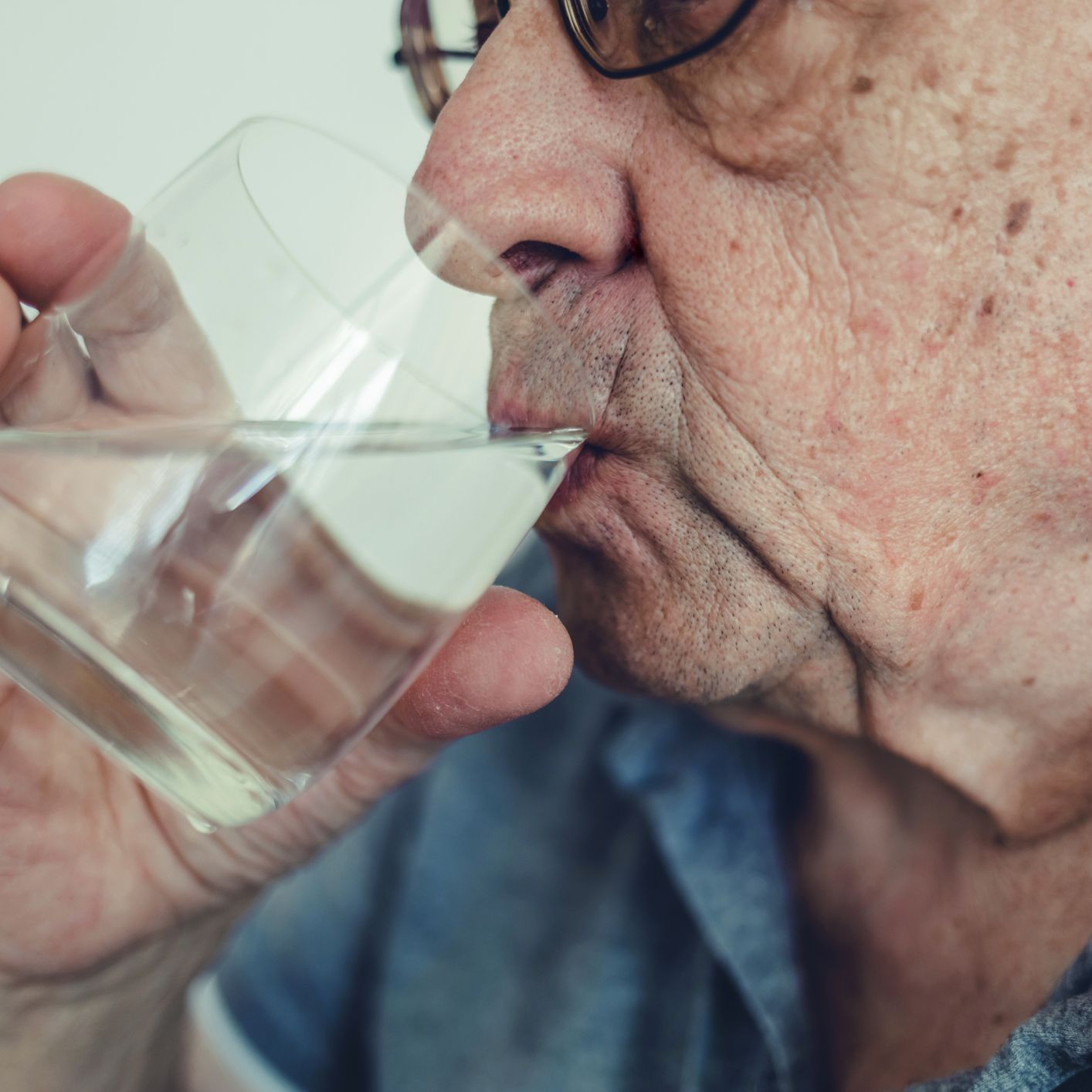 Close-up of an older person wearing glasses drinking water from a clear glass.